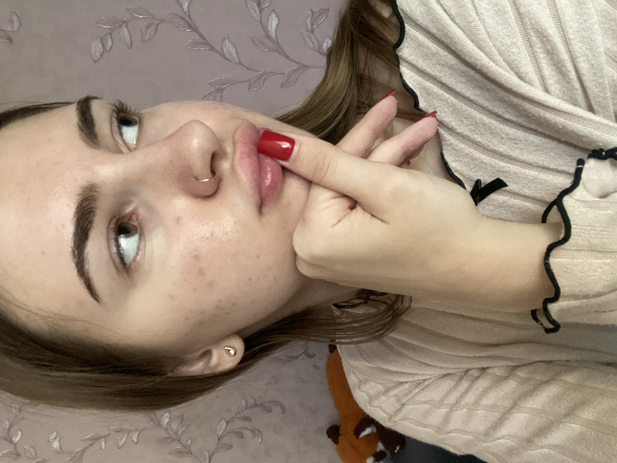 A medium shot captures a young woman with light brown hair, adorned with a small gold earring and a nose ring. She is wearing a light pink ribbed sweater with black trim. Her right hand is raised to her chin, with her index finger touching her lips. Her nails are painted a vibrant red. The background is a pink wall with a faint floral pattern. The scene appears to be indoors, likely in a residential setting. There is no indication of the location being in Дьяково, Russia.