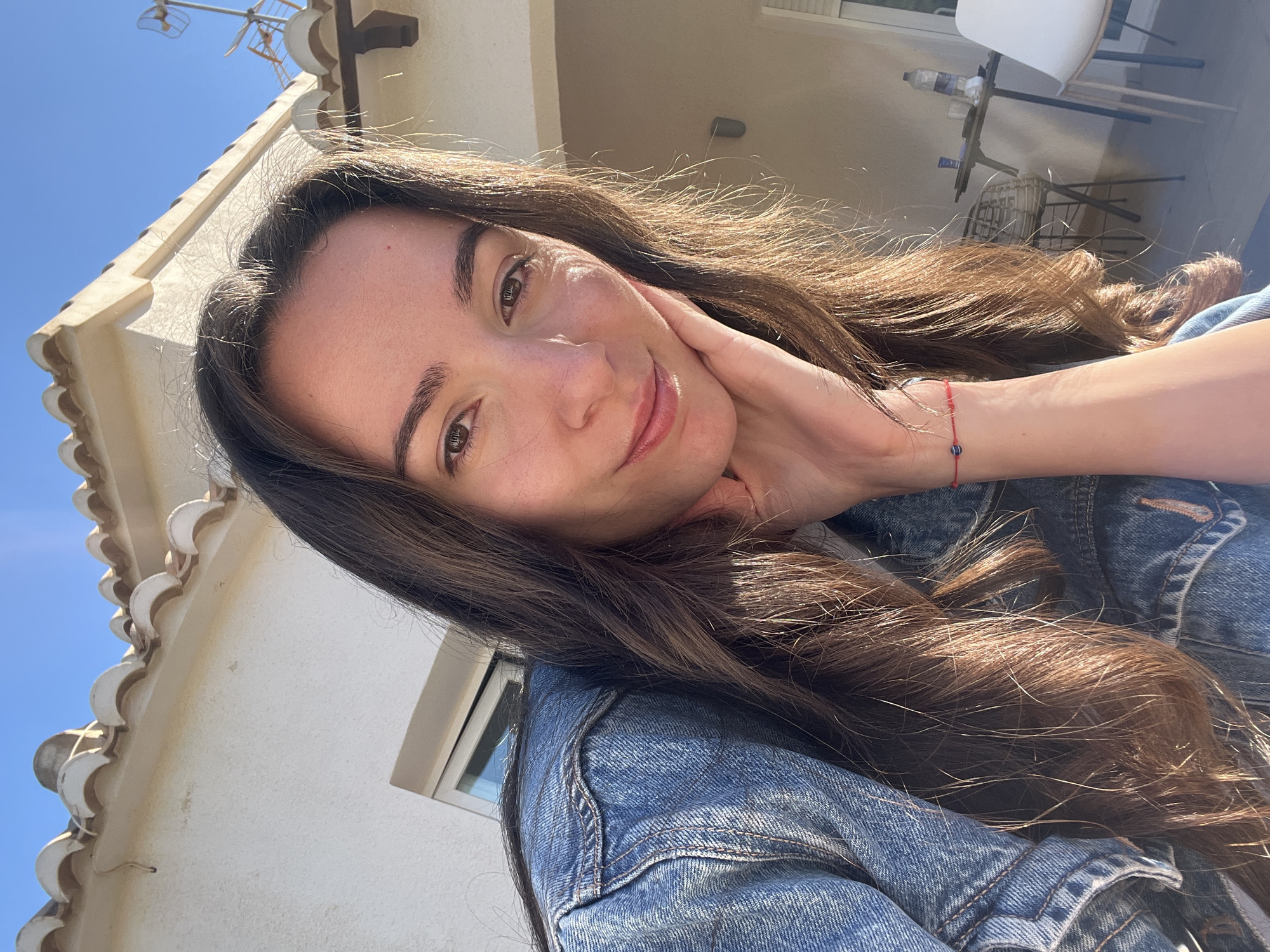 A young woman with long brown hair poses for a selfie. She is wearing a blue denim jacket and has a red bracelet on her wrist. Her right hand is resting on her face. The background shows a building with white walls and a tiled roof under a clear blue sky. In the distance, a table and chairs are visible. The location is likely el Poble Nou de Benitatxell / Benitachell, Spain, based on the architectural style.