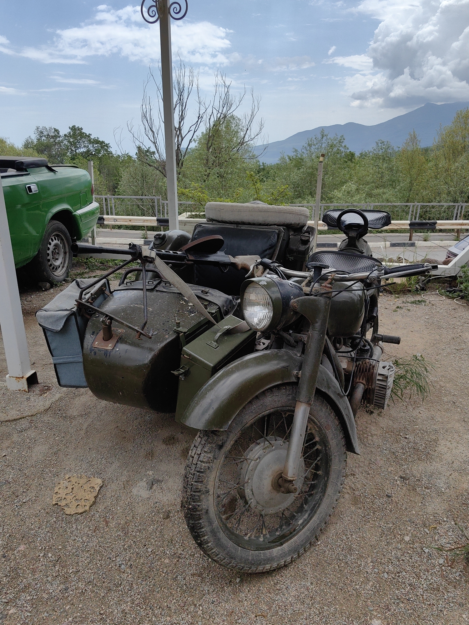The image presents a vintage military motorcycle with a sidecar, located in what appears to be an outdoor display or parking area. The motorcycle is painted in a dark olive green color and exhibits signs of wear and age. The tire in the front is worn, indicating some use. The sidecar is attached to the left of the motorcycle, and it also shows its age.
The surrounding environment includes a gravelly ground surface, some vegetation, and parts of a green vehicle. The sky is partly cloudy, and mountains are visible in the background. Based on the prompt, the location is Luchistoe, Ukraine.