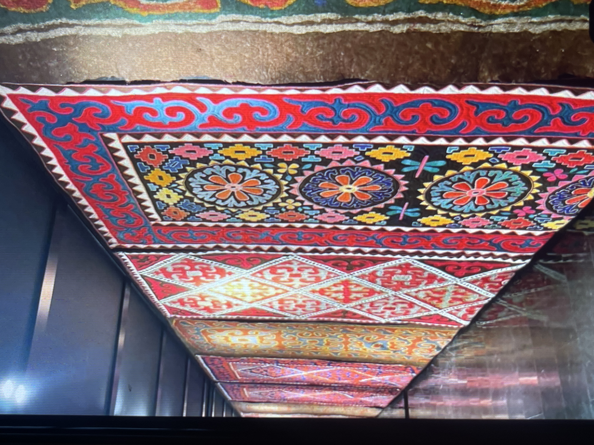 The image depicts a display of vibrantly colored textiles, likely rugs or wall hangings, arranged vertically. The foreground textile features intricate patterns with floral motifs in shades of red, blue, yellow, and pink against a black background, bordered by a red and blue scroll design and a zig-zag pattern. Behind it, several other textiles are visible, displaying diamond shapes and geometric patterns in warm tones of red, orange, and yellow. The textiles appear to be displayed indoors, possibly in a market or a museum, with a dark ceiling overhead and a reflective floor surface. A portion of a green textile with colorful embroidery is visible on the far right. The scene conveys a rich cultural display of textile art. This type of textile display is commonly found in traditional bazaars and cultural centers within Bishkek City, Kyrgyzstan.