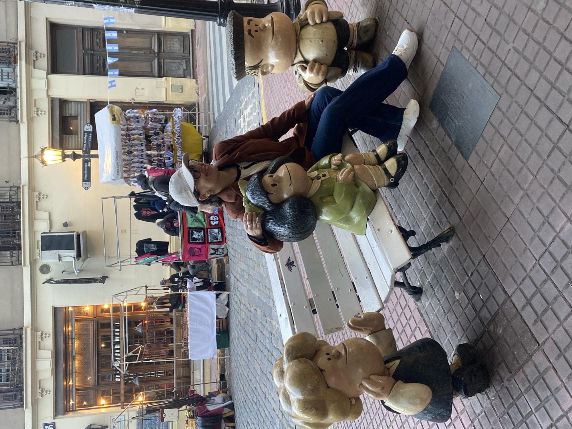 A woman in a brown jacket, blue jeans, and white sneakers and a white baseball cap is reclining on a bench between cartoon character statues: Mafalda, Manolito, and Felipe. She appears to be resting. The statues are part of a display located in the San Telmo neighborhood of Buenos Aires, Argentina, specifically on Defensa Street. The street is lined with shops and vendors, and Argentine flags are visible. The walkway is paved with red and gray bricks.