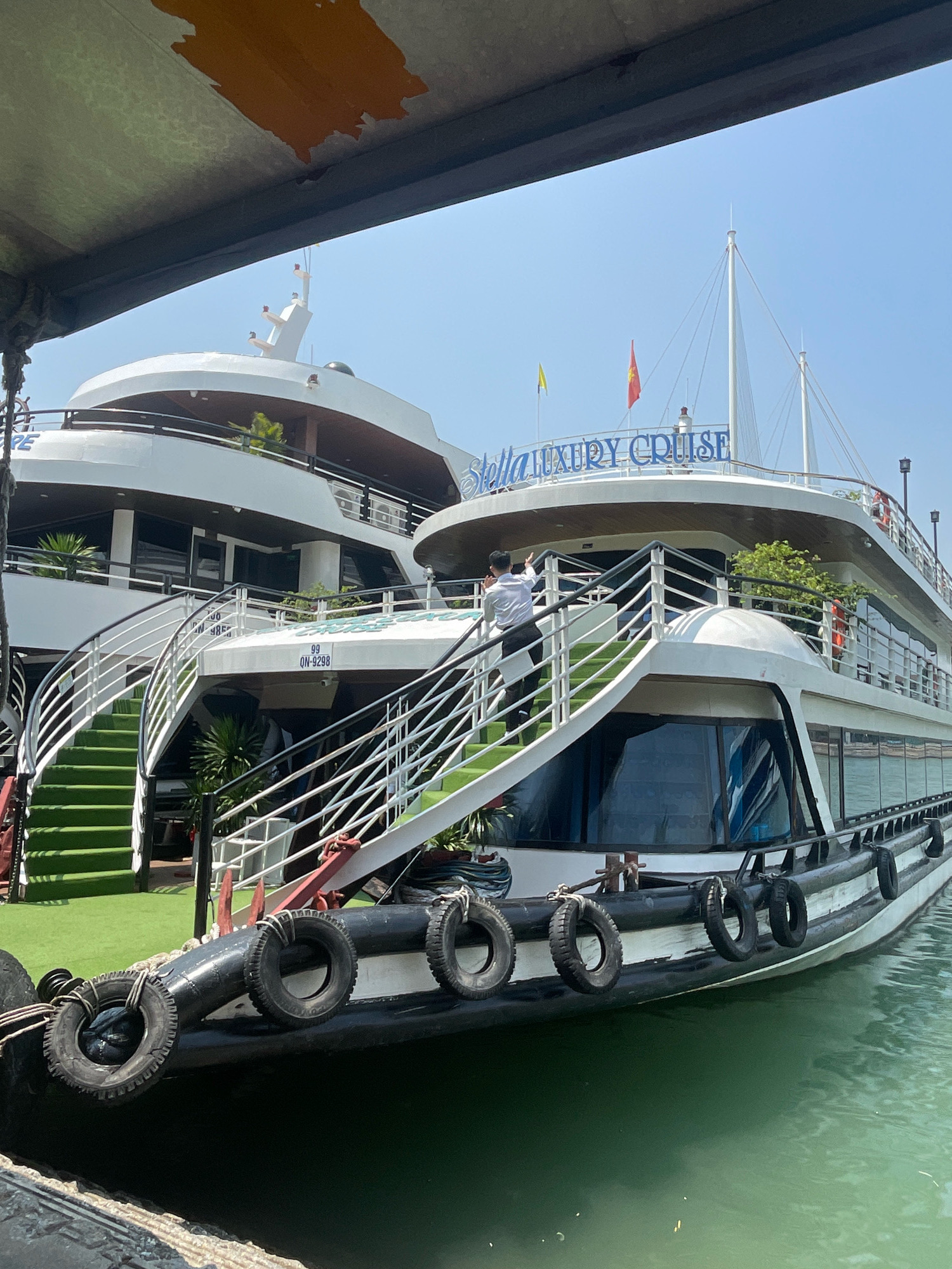 The image shows a large white luxury cruise ship docked in the waters of Ha Long Bay, Quang Ninh City, Vietnam. The ship, named "Stella LUXURY CRUISE" in blue lettering, has multiple decks, white railings, and large windows. Two flags, one yellow and one red with a star, fly from poles on the upper deck. Several people are visible on the ship, including one in a white shirt and black pants waving from a staircase. The staircase is covered with green artificial turf. The hull of the ship is black and white, with black tires acting as fenders. The water in the foreground is a murky green color. The sky is clear and blue. The image is taken from another vessel, partially obscuring the top of the frame with a dark overhang.