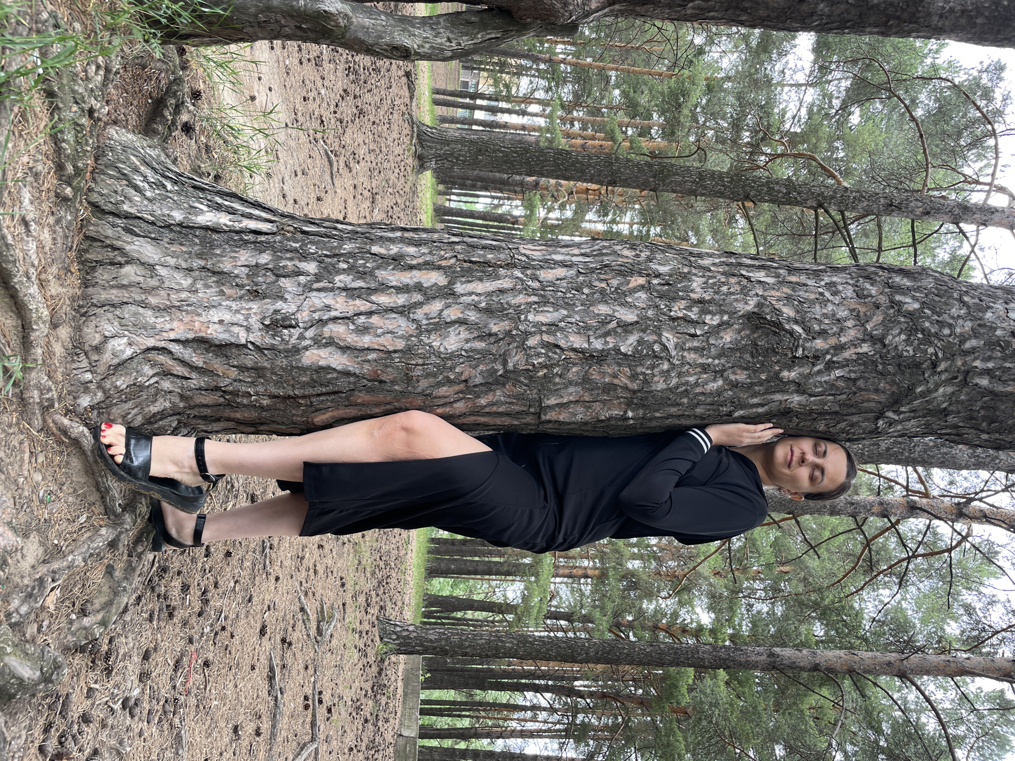 A young woman in a black dress leans against a large pine tree in a forest. She has her eyes closed and her hand resting on the tree trunk. The forest floor is covered in pine needles and cones. In the background, more pine trees are visible, and a building can be seen in the distance. The scene is likely located in a park or natural area within the city of Chaikovsky, Russia, given the prevalence of pine forests in the region.