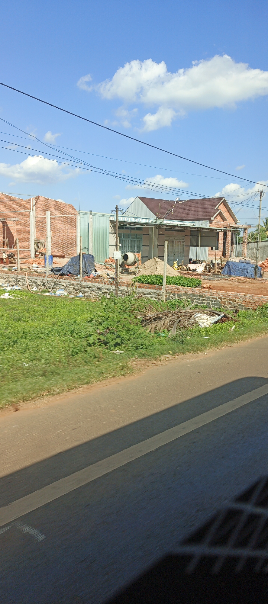 The image shows a construction site on a sunny day in Chư Sê, Vietnam. In the background, there are two houses under construction, one with brick walls and another with a completed brown roof. Construction materials such as bricks, sand, and tarps are visible. A cement mixer is present, and there are piles of sand and green plants. The site is bordered by a road in the foreground, with a patch of grass and vegetation to the left. Power lines are strung across the bright blue sky with scattered white clouds. The scene depicts typical rural construction activity.