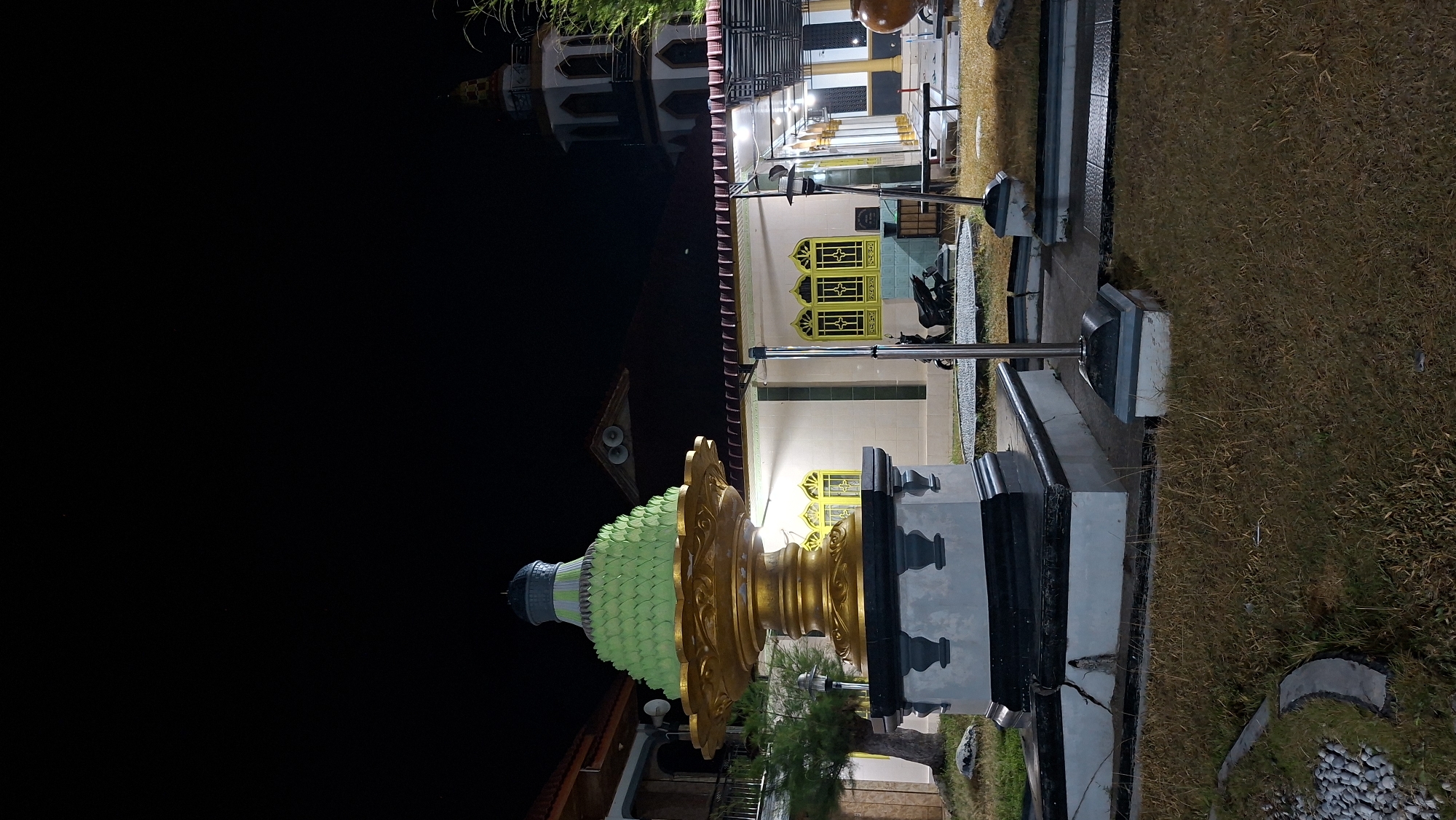 The image depicts a night scene in Lhokdagang, Indonesia, featuring a large, ornate golden fountain-like structure with a layered, green, conical top in the foreground. The fountain is situated on a multi-tiered, dark-colored base with grey accents. Behind the fountain, a mosque with a tall minaret is visible, illuminated by artificial lights. The mosque has a white facade and distinctive yellow-framed windows. The surrounding area appears to be a courtyard or garden, with dry grass, a few trees, and what looks like a paved pathway. A motorcycle is parked near the mosque, and outdoor seating or tables are also present under a covered area. The sky is dark, indicating it is nighttime.
