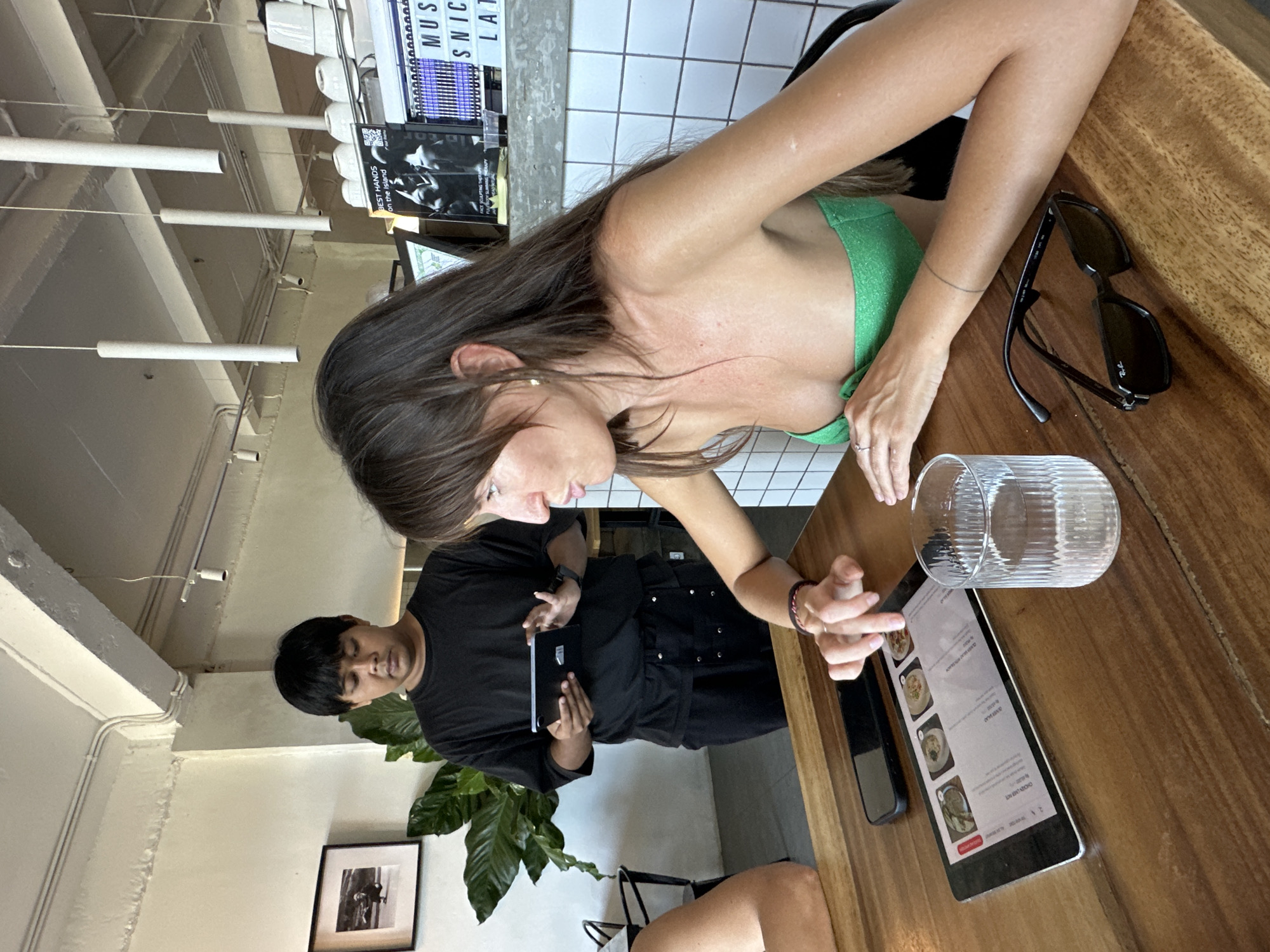 The image depicts a scene at an establishment in Kuta Selatan, Indonesia. A woman in a green top is seated at a wooden table, interacting with a tablet showing a menu. Her finger is pointing towards the screen. A glass of water is nearby. On the table, there is a pair of sunglasses and a cell phone. A male server wearing a black uniform stands near the table, holding a black notepad. A potted plant is visible in the background. A framed photograph is mounted on the wall. The interior has white walls and tiled sections. Overhead lights are suspended from the ceiling. There are also posters on the wall.