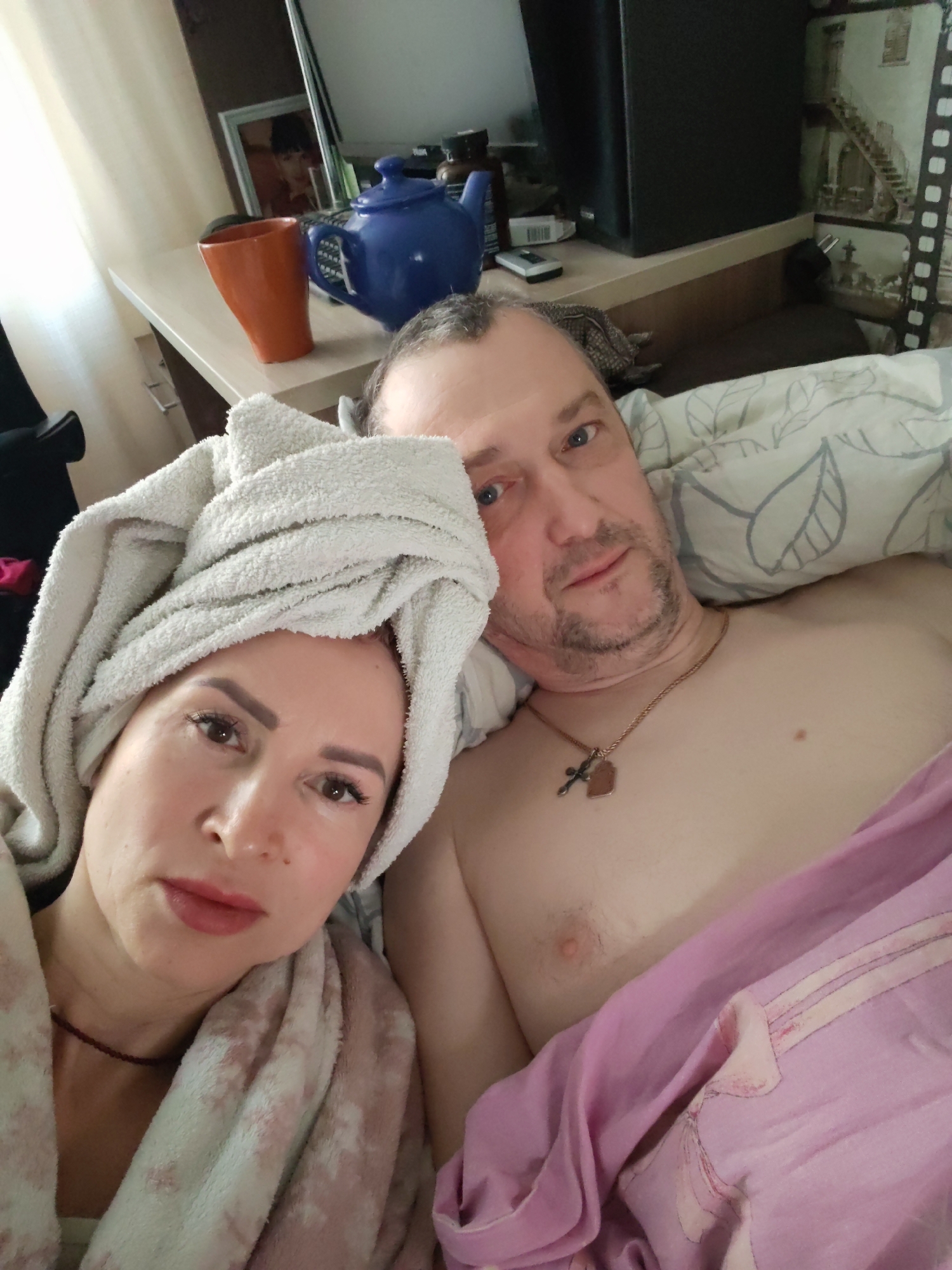 A selfie shows a middle-aged couple lying in bed, possibly at a residence in Chaikovsky, Russia. The woman is on the left with a white towel wrapped around her head and a light-colored robe. She is looking directly at the camera. The man is lying to her right, shirtless under a pink blanket, with a silver chain and cross necklace visible. He is also looking at the camera. A bedside table is visible in the background, cluttered with a mug, a blue teapot, and other items. The wallpaper behind the table features a vertical filmstrip design with images of architecture.