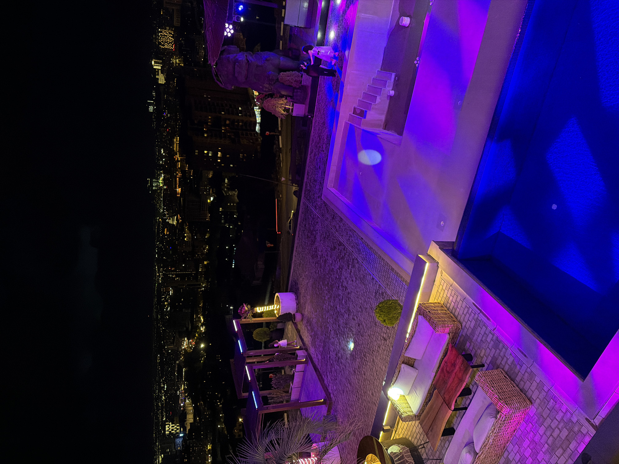 This image depicts a vibrant rooftop lounge or terrace at night, offering a panoramic view of a city illuminated by lights. The scene is characterized by dynamic artificial lighting, predominantly in shades of purple and blue, creating a contemporary and festive atmosphere.
In the foreground and midground, a sophisticated outdoor area is laid out. A rectangular swimming pool, brightly lit with blue underwater lights, is a central feature. Adjacent to the pool, comfortable seating arrangements are visible, comprising wicker couches and chairs with white cushions, placed on a paved, cobblestone-like surface. These seating areas are accented with warm yellow and white strip lighting, along with spherical globe lights. Several pergola-like structures with integrated lighting contribute to the venue's design, and various potted plants, including a palm and smaller bushes, are strategically placed, some adorned with decorative lights.
Towards the left side of the terrace, a large, dark statue, resembling an ape or a similar imposing creature, stands prominently. Near this statue, a few individuals are gathered, appearing to be socializing; one person is seen in lighter attire, and another in darker clothing. Other people are intermittently visible across the terrace, suggesting a social event or an evening out.
The background unveils a vast cityscape extending into the distance, with countless building lights twinkling against the dark night sky. The overall impression is that of an upscale and lively venue designed for evening entertainment and relaxation. While not explicitly visible in the image, the provided context indicates the location is Yerevan, Armenia. No discernible text is visible within the media.