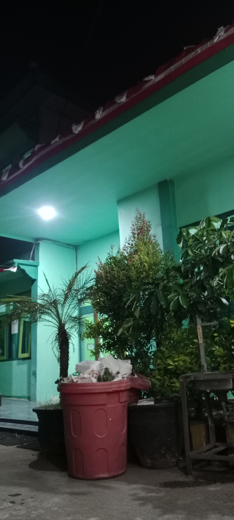 A medium shot shows a dark exterior scene at night in Tuban, Indonesia, featuring a building with a teal-colored facade and a red-trimmed awning. A bright overhead light illuminates a section of the building's exterior. In the foreground, a large red trash bin overflows with white plastic bags. To the left of the bin is a small palm tree in a pot, and to the right are several larger potted plants, including a bush with red-tipped leaves and a plant with broad, dark green leaves. A dark metal structure, possibly a table or shelving unit, is visible behind the potted plants. The ground is a concrete surface.