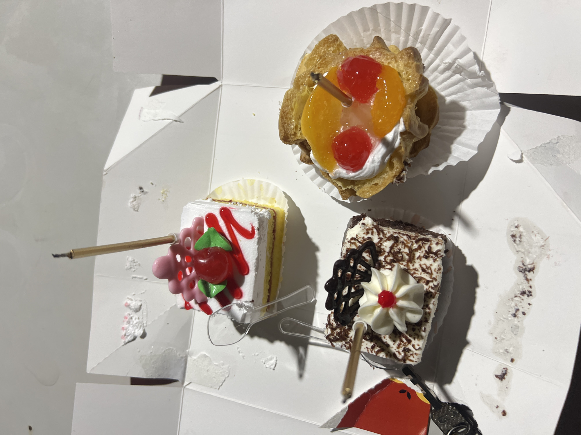 The image shows three small cakes in a white cardboard box. One cake is square with white frosting, yellow layers, and red icing swirls, topped with a pink cherry and green leaves. Another cake is rectangular, with dark frosting and shredded chocolate, topped with white frosting, a red cherry, and chocolate swirls. The third is a round pastry with cream, sliced peaches, and two red cherries on top. All cakes have candles. The box appears to be a takeout container. The setting is indoors, with artificial lighting casting shadows. There are no people visible. This image was taken in Denpasar, Indonesia.