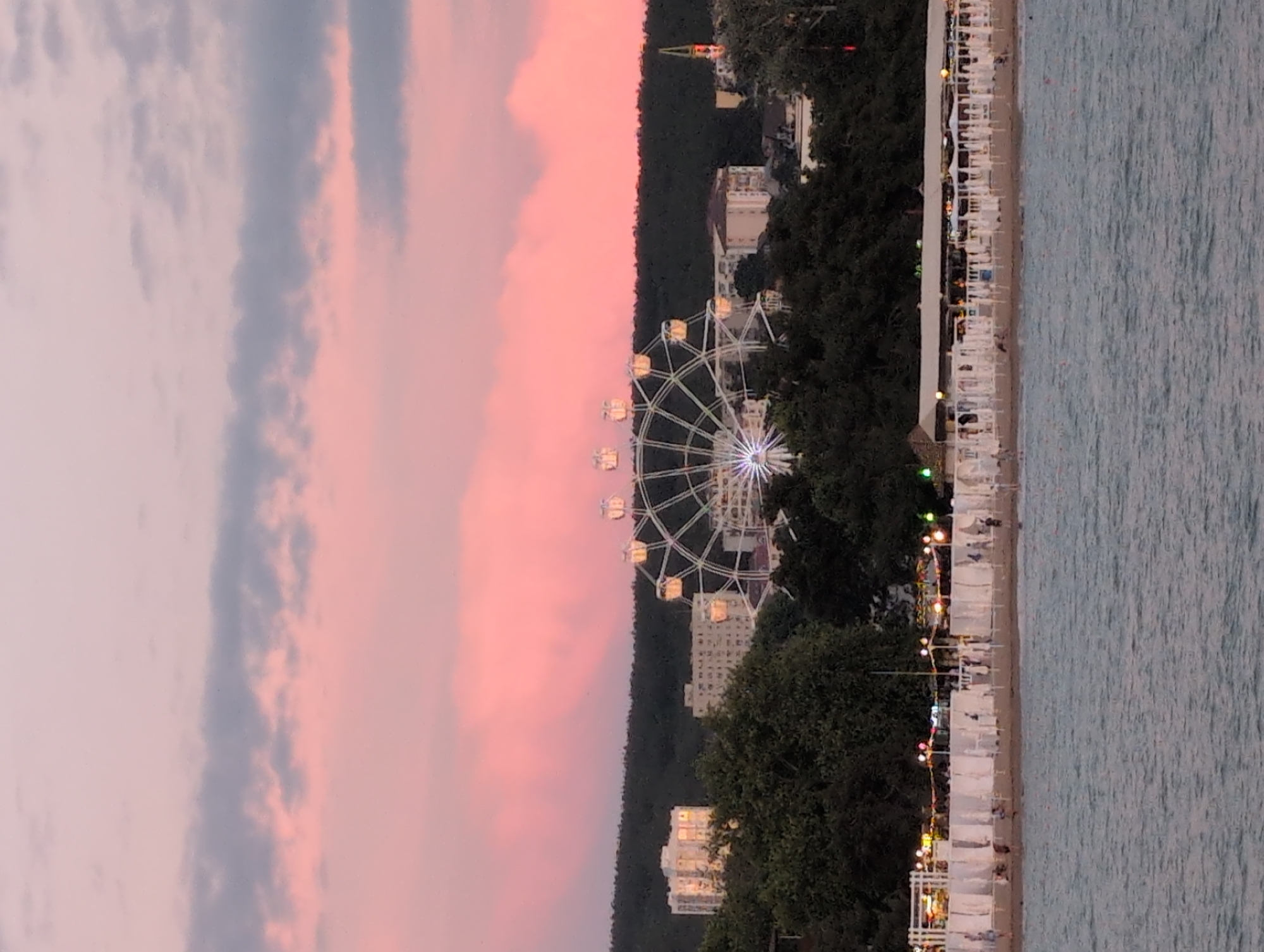 This image depicts a panoramic view of the Gelendzhik embankment and bay at dusk. In the foreground, the calm waters of the bay are visible, with ripples on the surface. Beyond the water, a sandy beach stretches across the frame, lined with white beach umbrellas and small seating areas. People are scattered across the beach, engaged in various leisurely activities. Above the beach, a line of trees separates the embankment from the city skyline. In the background, several multi-story buildings rise above the treeline, some appearing to be residential or hotel complexes. Prominently featured in the mid-ground is a large Ferris wheel, illuminated with white lights, its spokes and cabins clearly visible against the colorful twilight sky. The sky itself is a gradient of soft pink and lavender hues, indicative of a sunset or sunrise. The overall scene conveys a peaceful and vibrant atmosphere of a seaside resort town.