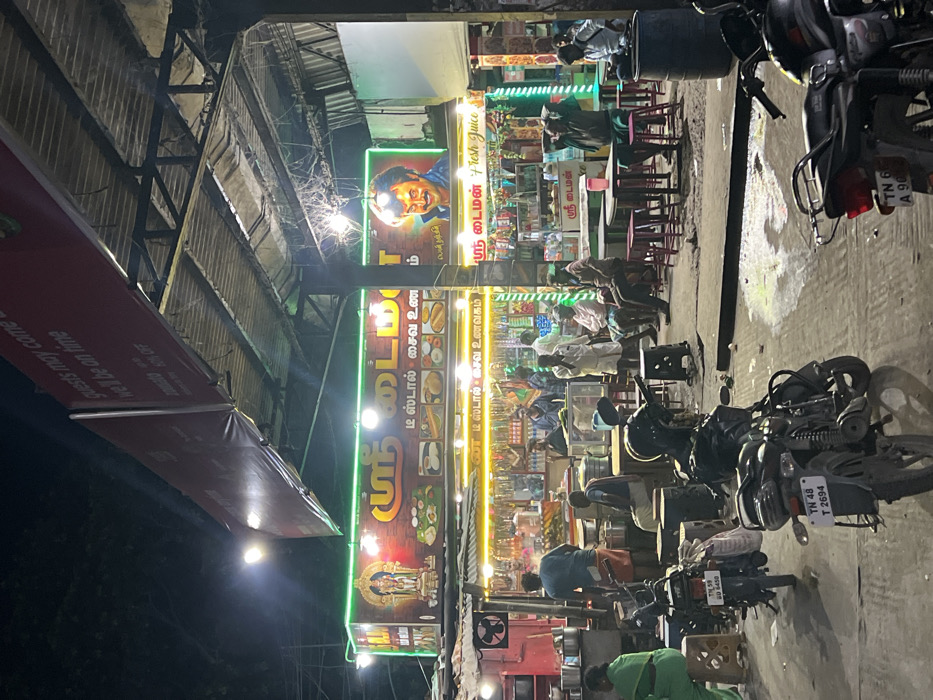 This image captures a vibrant nighttime scene at a street-side food establishment in Palani, India. The setting is brightly illuminated by artificial lights, including neon green and yellow strips outlining prominent signs.
Numerous people are present, indicating a busy and popular spot. Some individuals are seated at small tables with stools, appearing to dine or socialize. Others are standing near the counters or in the general area. The activity suggests a casual dining or takeaway experience.
The establishment consists of several adjoining stalls. Large vertical signs feature Tamil script, prominently displaying "ஸ்ரீ டைம்" (Sri Time) and "டீ ஸ்டால் சைவ உண" (Tea Stall Vegetarian Food). One sign further clarifies "ம் ன டீ ஸ்டால் சைவ உணவகம்" (Tea Stall Vegetarian Restaurant). A separate banner advertises "ஸ்ரீ டைமன் Presh Juice" (Sri Daimon Fresh Juice). A large portrait of a man, characteristic of regional public figures, is displayed on one sign, while another shows an ornate image resembling a deity or temple entrance.
Several motorcycles are parked on the paved ground, with visible Indian license plates such as "TN 48 T 2694", "T 59 206450" (partially obscured), and "TN 6 A 90" (partially visible), affirming the location in Tamil Nadu. Above the stalls, a corrugated metal roof structure is visible. On the left, a red awning extends, bearing partially visible English text: "guests may come well be on time". The overall atmosphere is lively and typical of an Indian urban street food scene after dark.