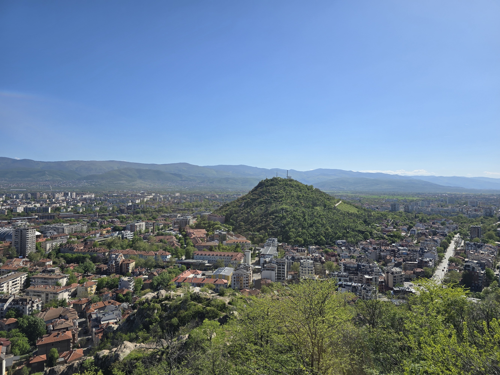 The image captures a panoramic view of a city, possibly Plovdiv, Bulgaria, dominated by a prominent green hill in the center. This hill, which is likely one of the city's iconic landmarks, has communications towers on top. Buildings of varying heights and architectural styles spread across the landscape surrounding the hill. In the distance, a range of mountains stretches horizontally. The sky is a clear blue, indicating a sunny day. Trees and greenery are visible in the foreground, adding depth to the scene.