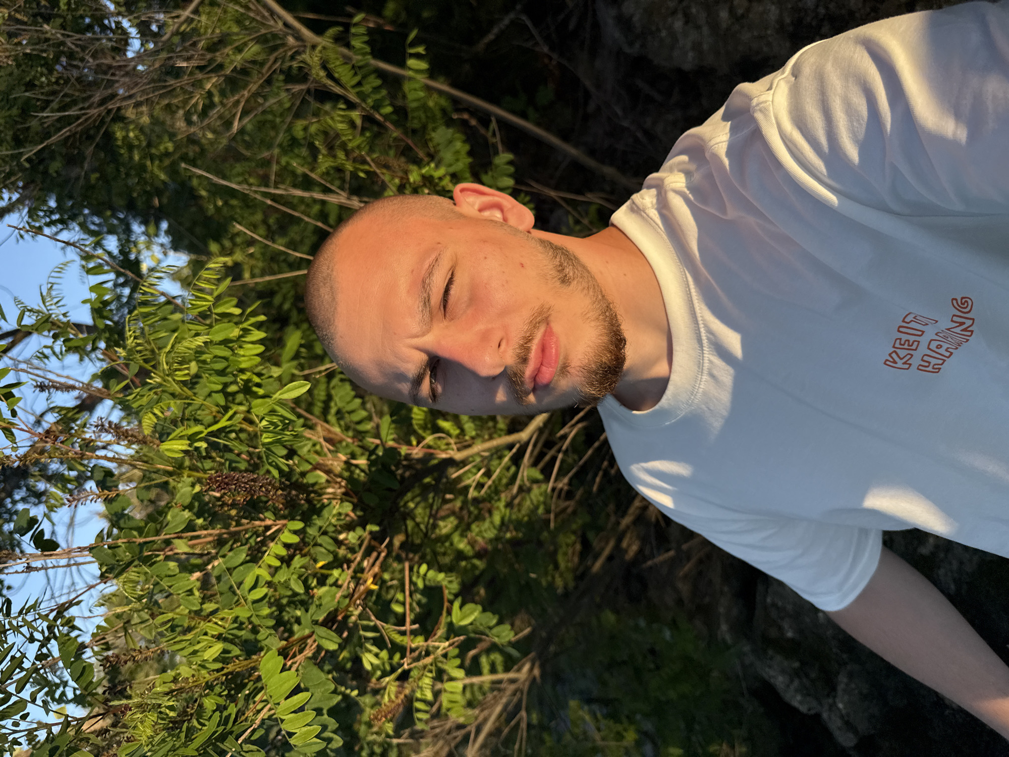 A young man with a shaved head and a goatee is taking a selfie outdoors. He is wearing a white t-shirt with orange text that appears to say "KEIT HANG". He is positioned in the foreground, with a backdrop of green foliage and trees. The lighting suggests it is either early morning or late afternoon, with the sun casting warm light on his face and shirt. Rocks are visible in the lower left corner of the image. The location is specified as Koprinka, Bulgaria.