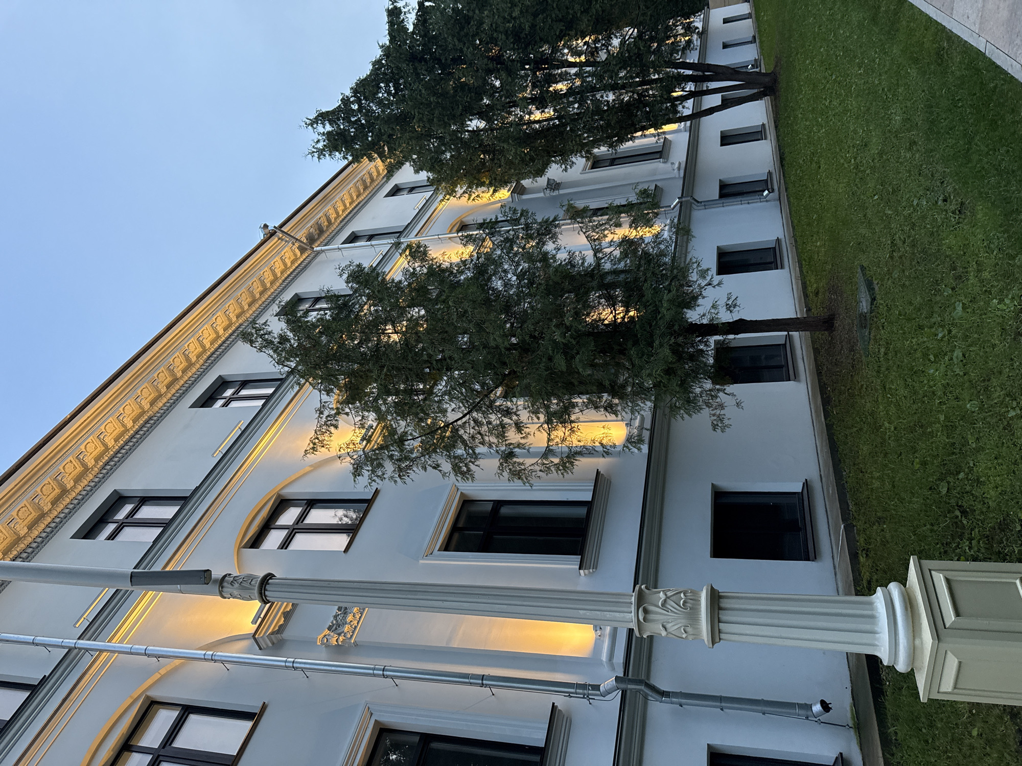 The image depicts the exterior of a multi-story white building with dark-framed windows, located in Moscow, Russia. The building features ornate architectural details along its upper facade, illuminated by warm yellow lights. Several evergreen trees are positioned in front of the building, with lush green grass covering the ground. A decorative lamppost with fluted detailing stands in the foreground to the left. The sky is a clear, pale blue, suggesting it is either early morning or late evening. There are no people visible in the scene.