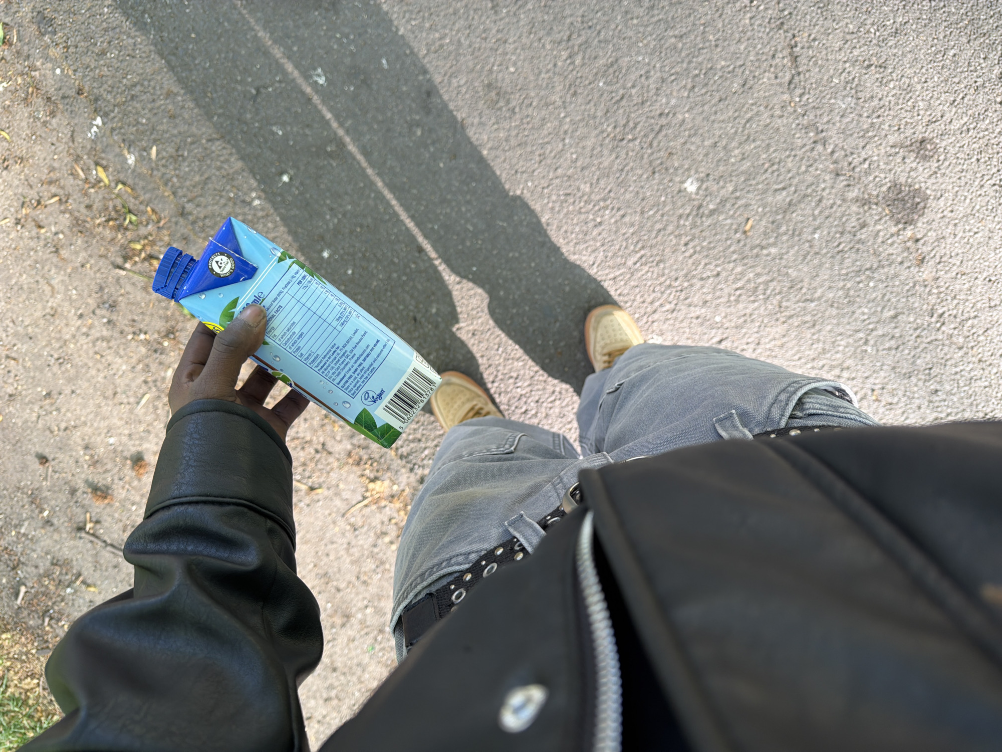The image is captured from a first-person, downward-looking perspective, showcasing the lower body of a person standing on an asphalt surface. The subject is wearing a black leather jacket, light-wash jeans, tan-colored shoes, and a black studded belt. In the left hand, a light blue carton of drink is held.
The scene depicts an outdoor setting on a bright, sunny day, evidenced by the long shadows cast across the rough asphalt pavement. Scattered dry leaves and small debris are visible on the ground.
The drink carton is light blue with a green leaf motif and water droplet graphics, featuring a blue screw-cap. Text on the carton identifies the brand as "Vita Coco" and displays comprehensive nutritional information, including values for energy, fat, carbohydrates (sugars), protein, salt, vitamin C, and potassium. The ingredients are listed as "Coconut Water (99%), Fructose." Important dietary information highlights that the product is "GLUTEN FREE, DAIRY FREE, SUITABLE FOR VEGAN," accompanied by a "Vegan" logo and a "PROTECTS WHAT'S GOOD" emblem. Production details state it is "Produced in Sri Lanka" for "All Market Europe Ltd." located in "London, United Kingdom," with a French distributor also mentioned. Storage instructions and a barcode are also visible.