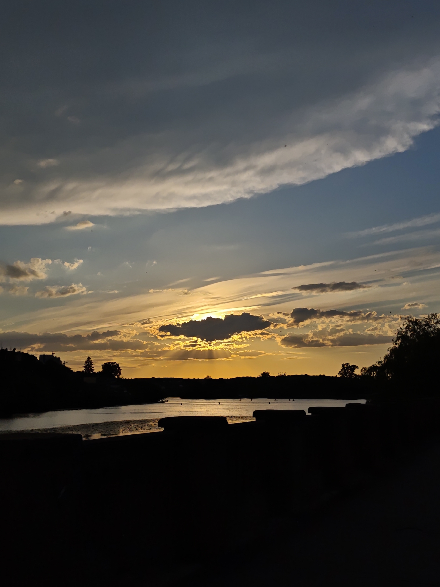 This media file captures a sunset over a body of water, likely a river or lake, in Vinnytsia, Ukraine. The sky is a dramatic display of colors, with streaks of orange, yellow, and blue intermingling with dark, heavy clouds. The sun is partially obscured by a dark cloud formation, casting a warm glow across the horizon and reflecting on the water's surface.
The foreground is dominated by a dark silhouette of what appears to be seating or a low wall along the water's edge, possibly a park or recreational area. Beyond this, a dark band of land represents the opposite shore, dotted with silhouetted trees and buildings on the left. The water itself is calm, with subtle ripples and reflections.
Several small, dark objects, possibly buoys or small boats, are visible on the water. The overall mood is serene and contemplative, typical of a sunset scene. The time of day is clearly evening, as indicated by the sunset lighting. The weather appears to be mixed, with both clear sky and cloud cover contributing to the atmospheric effect. No text is visible in the image.