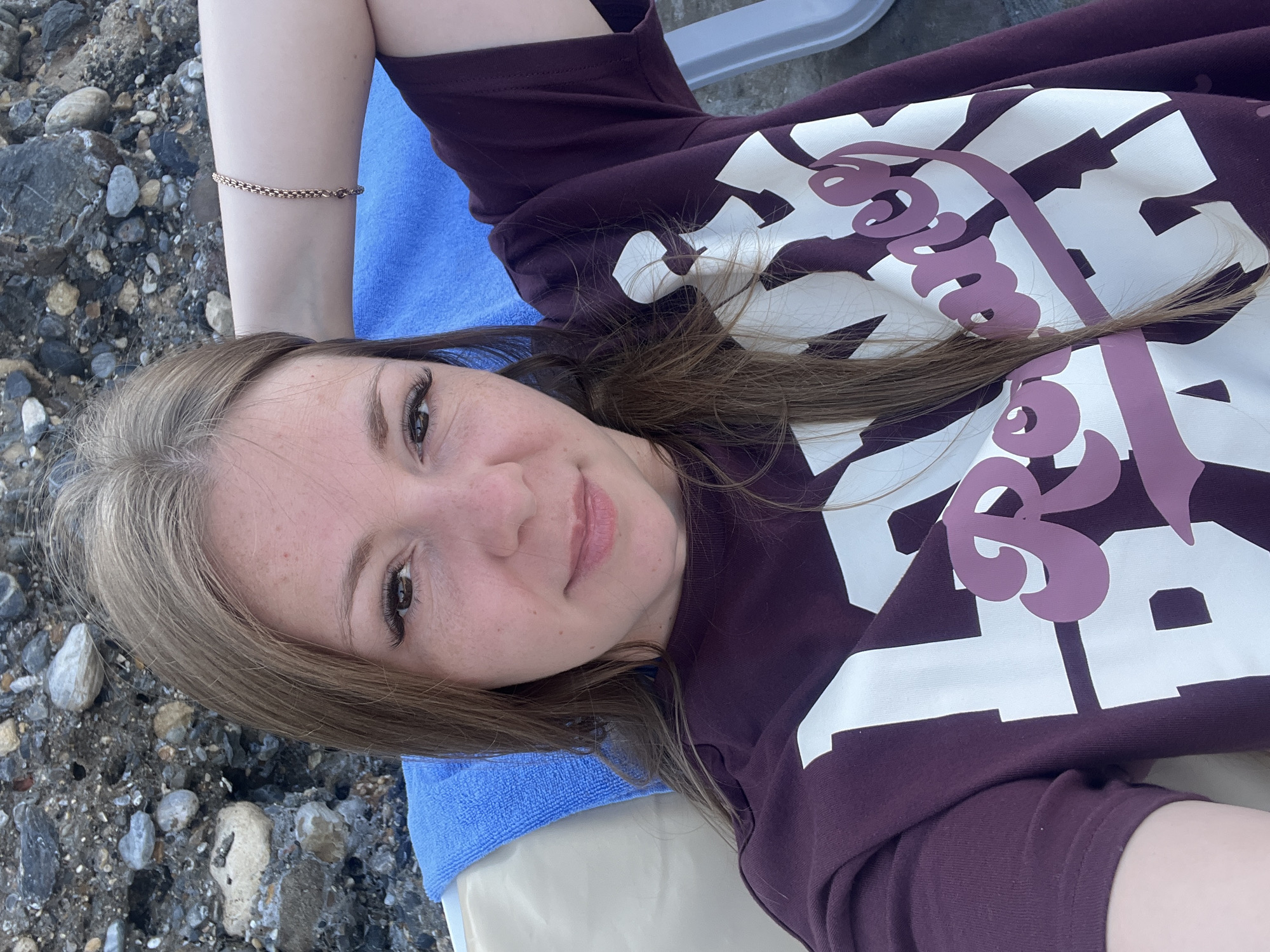 A young woman with long, light brown hair is lying on a beach chair. She is wearing a dark purple long-sleeved shirt with white and purple lettering that appears to say "Romance." Her right arm is propped behind her head, and she is wearing a delicate gold bracelet. A bright blue towel is draped behind her head. The background consists of a rocky shoreline. The image was taken from a selfie perspective. The scene is consistent with a beach environment in Alanya, Turkey, likely during daylight hours.