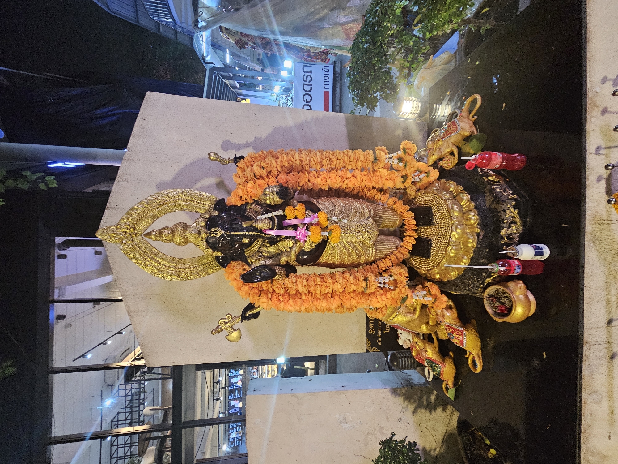 An outdoor shrine dedicated to the Hindu deity Ganesha is situated in front of a building with large windows in Bangkok, Thailand. The shrine features a large, dark-colored Ganesha statue adorned with an orange flower garland and a golden headdress. The statue is positioned on a black base surrounded by smaller golden rat figures, which are the vahana (mount) of Ganesha. Offerings, including incense sticks in a golden pot and small bottles of liquid, are visible at the base of the shrine. The background includes the building's interior visible through the windows, suggesting the shrine is part of a commercial or public space. The overall scene is illuminated by artificial lights, indicating it is nighttime.