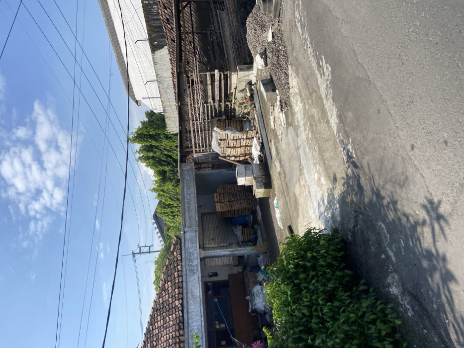 The scene shows a dirt driveway leading to a paved street in Mojosari, Indonesia. On the left side of the driveway, there is a residential building with a terracotta roof. The building has a concrete foundation and a white painted wall. There are several wooden pallets stacked against the building, and a small bush in the foreground. On the right side of the driveway, there is a wooden structure with a slatted roof and a white painted wall. Several wooden pallets are stacked in front of the structure. The driveway is littered with debris, including discarded plastic bags and rocks. There are many power lines overhead. The sky is blue with some clouds.