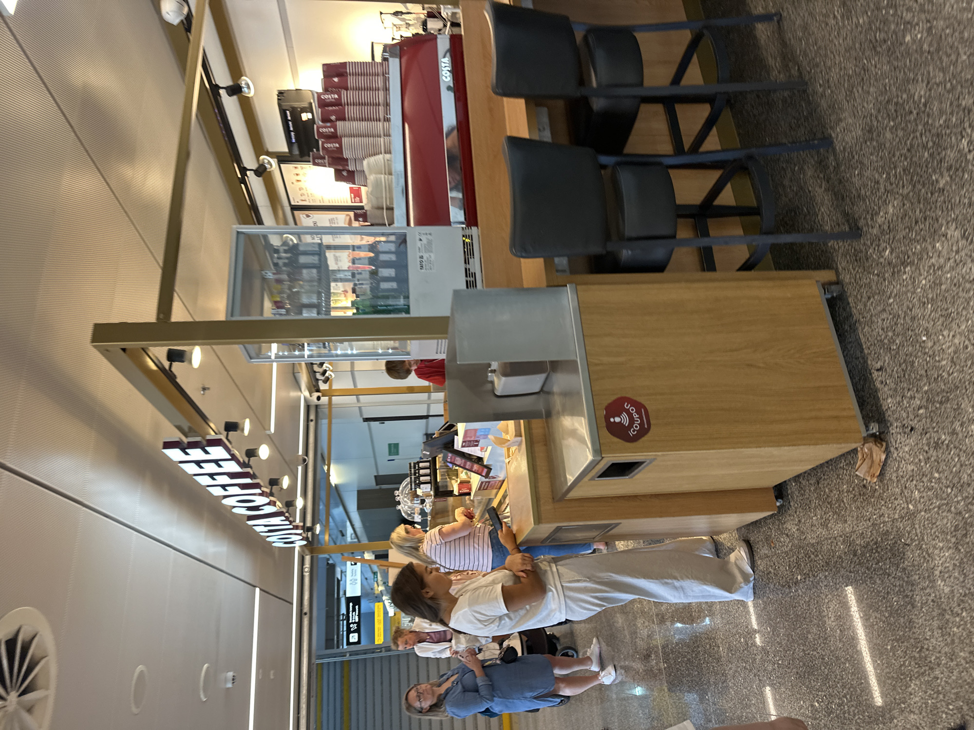 The image shows a Costa Coffee shop with a red espresso machine and stacks of coffee cups. A young woman in a white t-shirt and light-colored pants is standing at a counter, looking at her phone. She is holding a small, dark object, possibly a phone or card. Behind her, several other people are waiting in line. The shop appears to be located in an airport or train station, given the signage visible in the background. The overall scene is bustling, with customers interacting with the staff and each other.