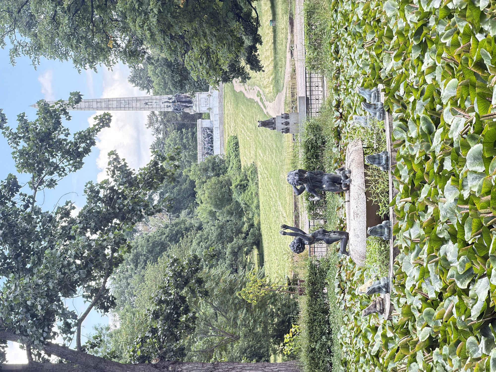 In Sofia, Bulgaria, the image captures a serene view of the Sofia National Military Cemetery, prominently featuring the Monument to the Soviet Army. In the foreground, a vibrant pond is filled with lush green lily pads. A circular stone fountain is situated in the pond, adorned with bronze sculptures of two children reaching upwards and several small frog statues positioned around its base. Beyond the pond, a winding path leads up a grassy hill towards the imposing Monument to the Soviet Army. This monument consists of a tall, obelisk-like structure topped with a star, and at its base are a group of bronze figures. The surrounding landscape is characterized by mature trees and manicured greenery, indicative of a well-maintained park or memorial site. The sky above is a clear blue with scattered white clouds.