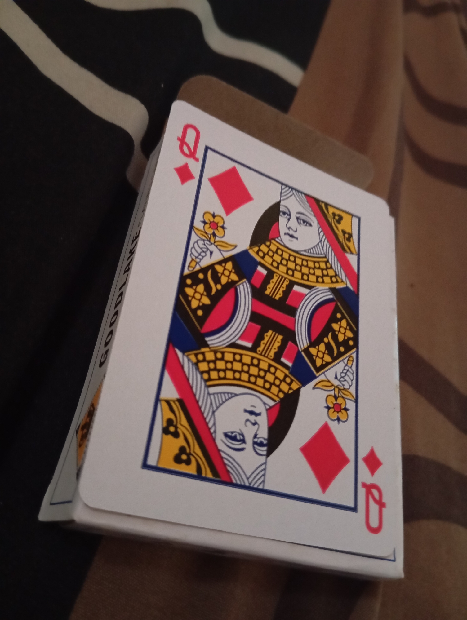 The image shows a close-up of a deck of playing cards, specifically featuring the Queen of Diamonds. The card is partially obscured by other cards in the deck and is propped up against something brown and textured, possibly a notebook or a box. The background consists of a fabric with a striped pattern in black and white, and another fabric with a brown, patterned texture. The playing card itself displays the traditional depiction of the Queen of Diamonds, with a red diamond symbol and the letter 'Q' visible. The text "GOODLAKE" is partially visible on the side of the deck. The scene is indoors, with soft lighting that creates shadows and highlights on the objects. There are no people visible in the image. The location in Sudikampiran, Indonesia, is not discernible from the visual content of the image.