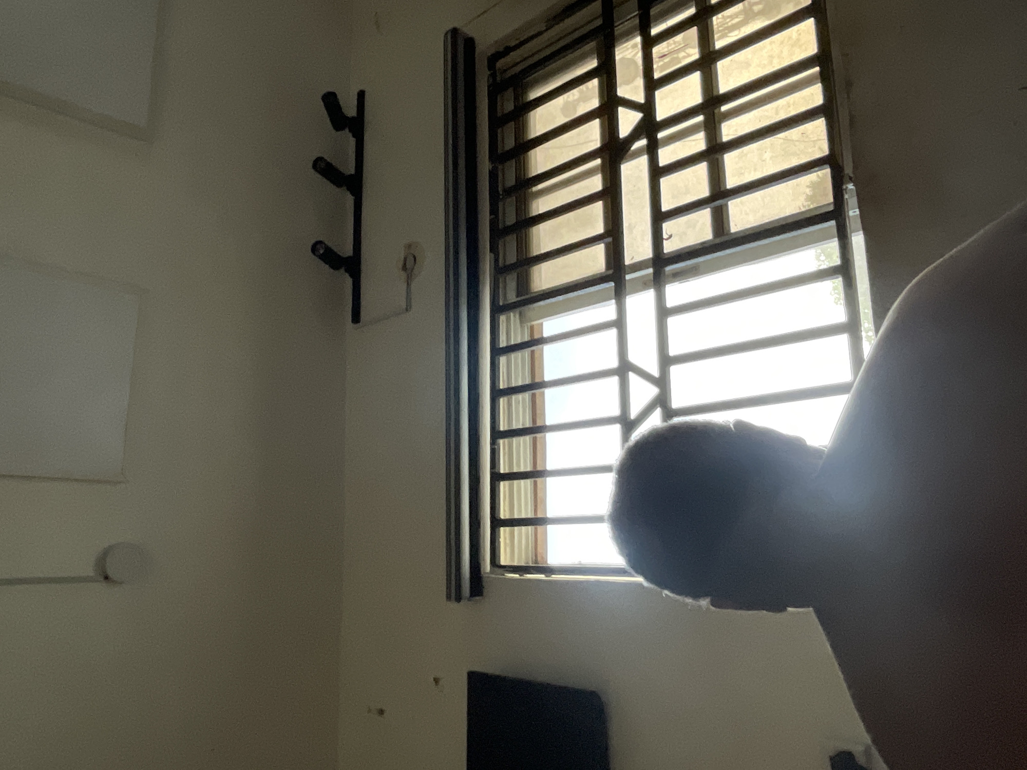 The image shows an interior shot with a window covered by a metal security grill, likely taken in Ado, Nigeria. A black metal coat rack is mounted on the wall to the left of the window. Part of a person is visible on the right side of the image, obscuring the view through the window. A black object, possibly a piece of furniture or equipment, is visible beneath the window. The walls appear to be painted in a light color.