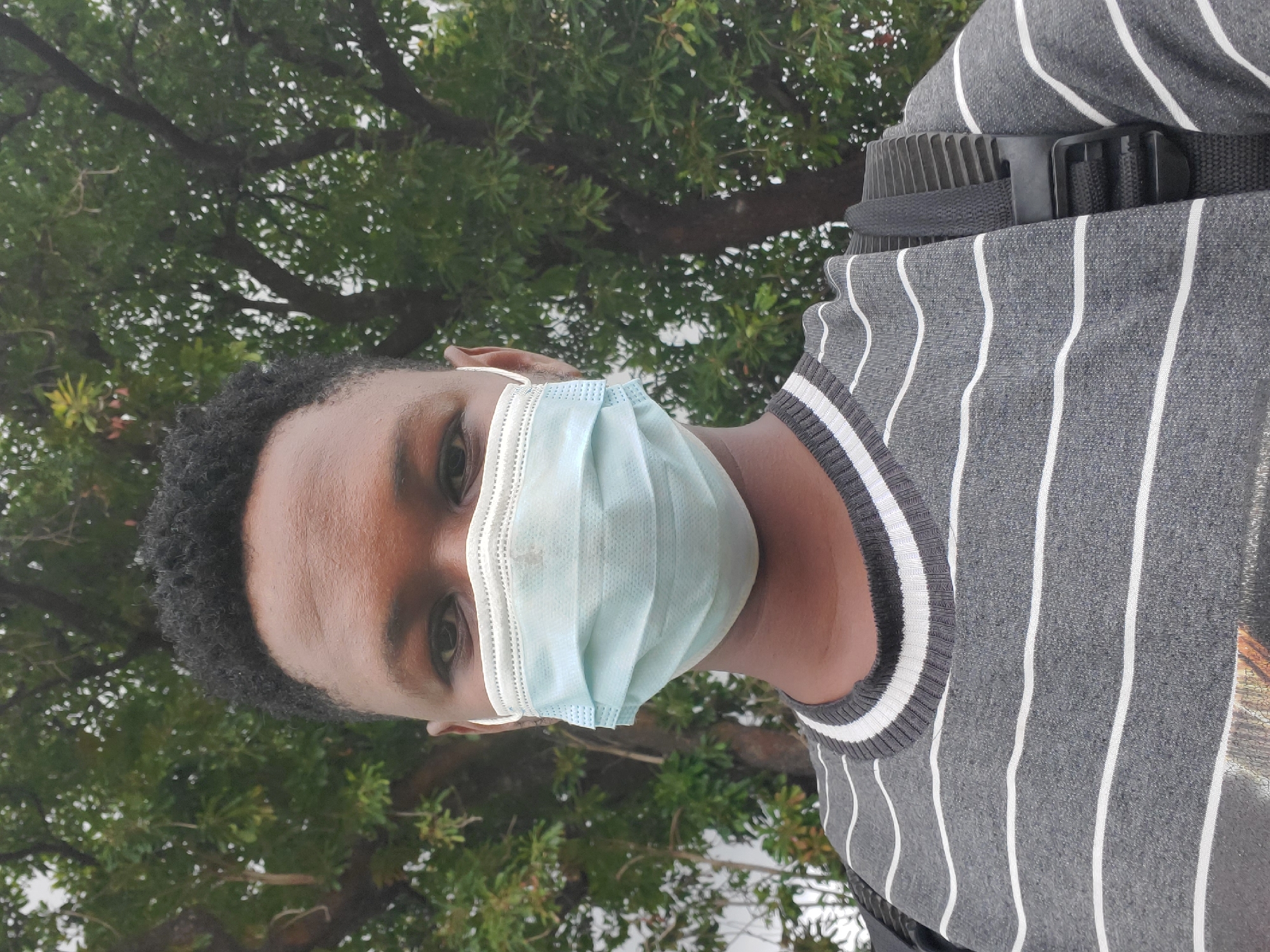 A person wearing a grey and white striped top and a blue surgical mask stands in front of a tree with green leaves. The background is slightly blurred. A black strap with a buckle is visible over their shoulder. The location is in the city of Mararaba, Nigeria.