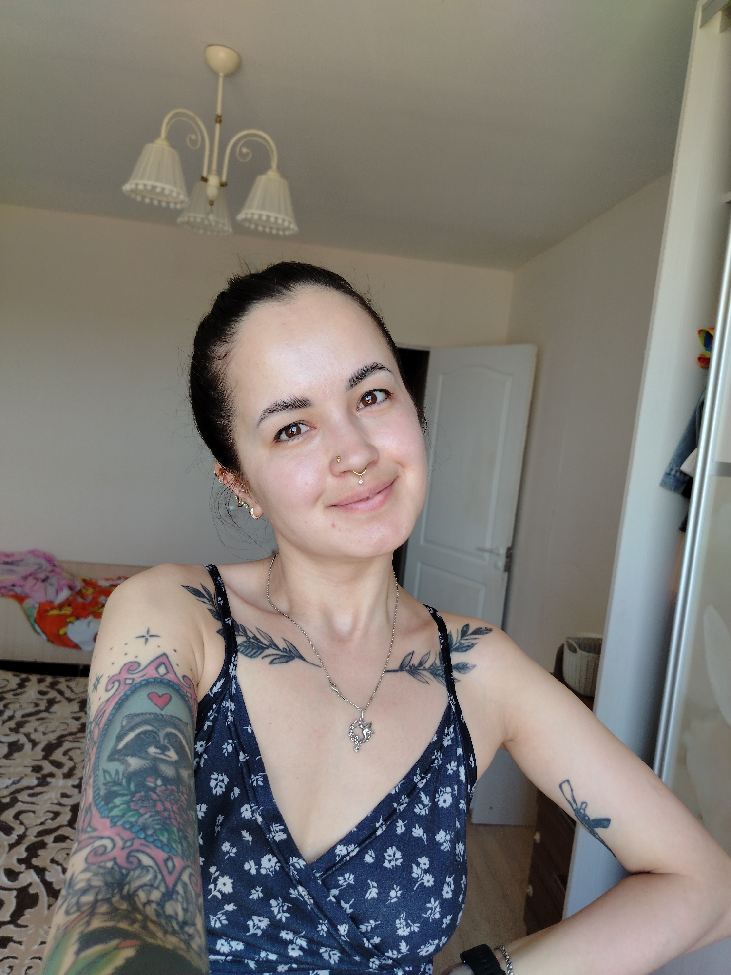 A young woman is taking a selfie indoors. She has dark hair pulled back, fair skin, and is wearing a floral-print sleeveless top. She has multiple tattoos on her arms and chest, including a raccoon tattoo on her left arm and a leafy design on her collarbone. She is wearing a nose ring and earrings. The background includes a bed, a door, a wardrobe, and a chandelier. There are no indications as to the city of Noviy, Russia.