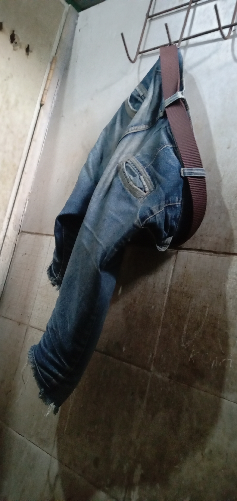 In a dimly lit room in Sumput, Indonesia, a pair of worn blue jeans hangs from a wire coat rack. A brown belt is looped through the jeans. The jeans show signs of wear and tear, with frayed edges at the bottom. The wall behind the jeans is tiled and marked with shadows, while the wall to the left appears to be painted and shows some damage. The scene is captured from a low angle, emphasizing the jeans hanging against the wall.