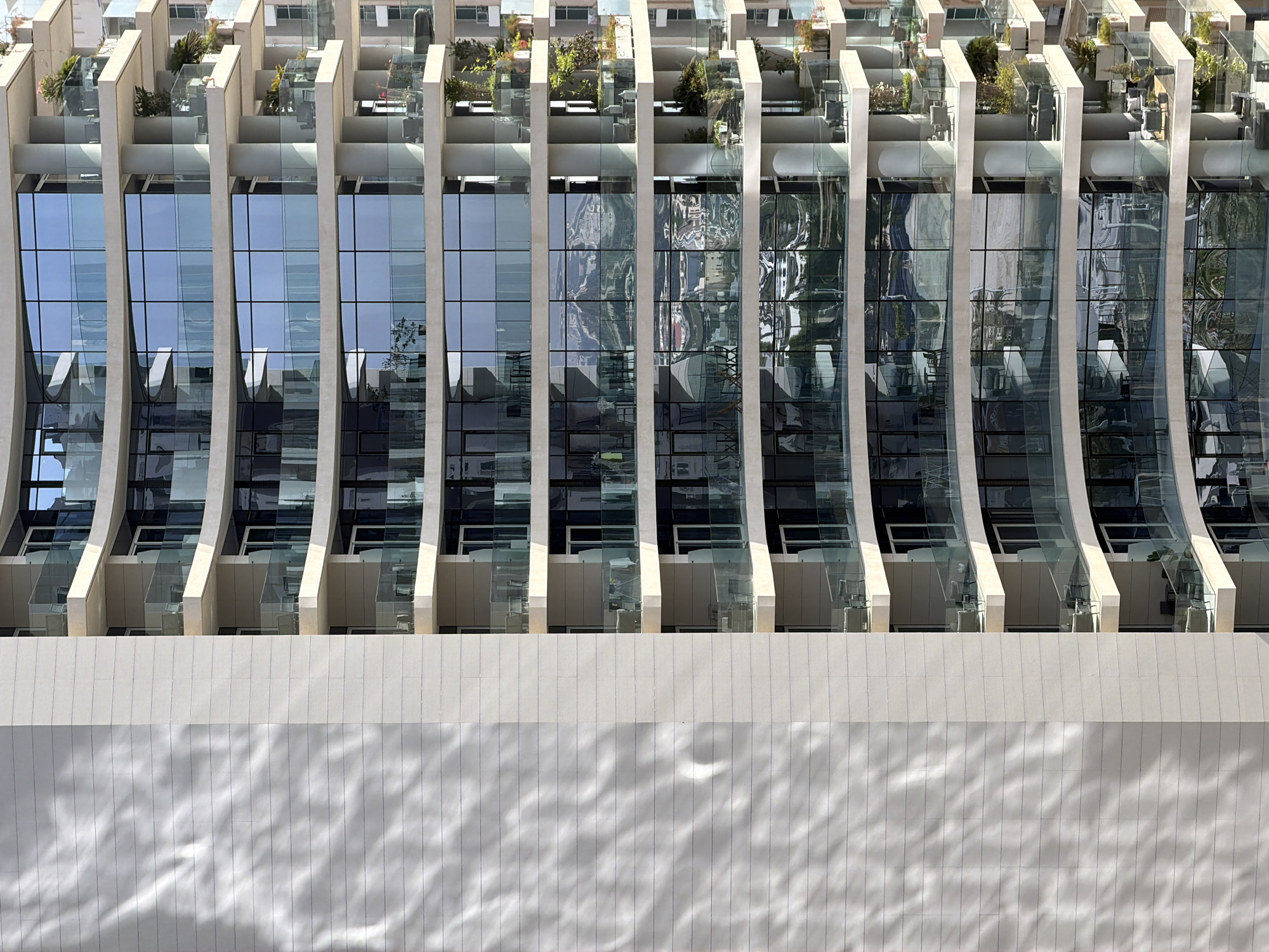 A close-up, high-angle, full-frame shot captures the exterior of a modern, multi-story building in an urban setting. The building features a textured, light-colored facade on the left, with horizontal lines and a dappled shadow pattern from what appears to be foliage. To the right, a series of balconies with glass railings overlook the city. These balconies are furnished with various seating arrangements and potted plants, some with lush greenery and others with more sparse vegetation. The building's facade is dominated by large, floor-to-ceiling windows that reflect the sky and surrounding landscape, creating a sense of depth and mirroring the urban environment. The windows are divided into rectangular panes, with some reflecting a distant cityscape and mountains. The building's architecture is characterized by curved lines and a tiered structure, with each floor slightly set back from the one below. The overall impression is one of contemporary urban living, with a focus on natural light and outdoor spaces.