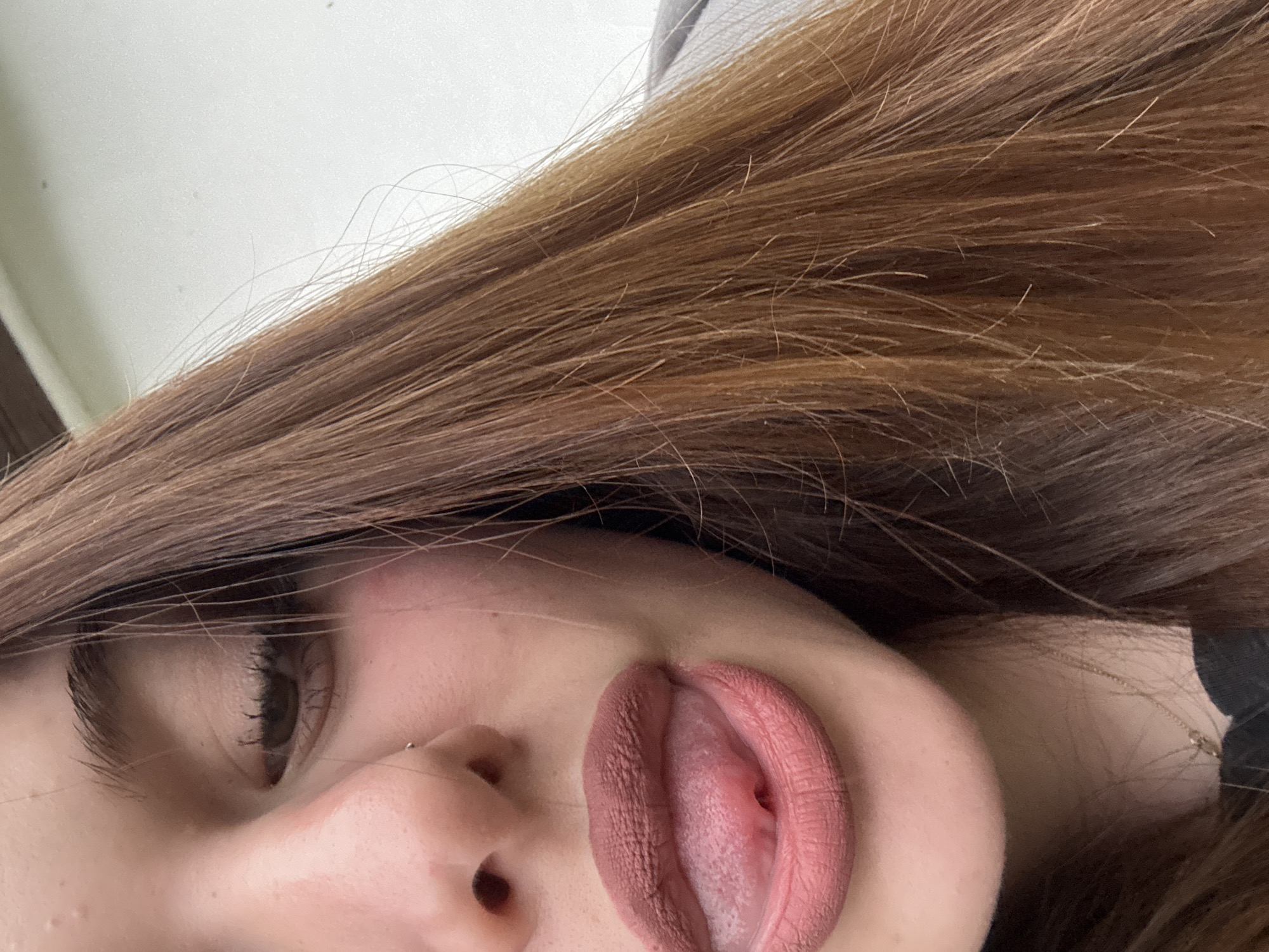 A close-up, angled view captures a portion of a person's face. Visible features include a brown eye with eyelashes, a nose with a silver stud in the nostril, and a mouth with the tongue sticking out. The lips are covered in a matte, nude-pink lipstick. Brown hair frames the face. Part of a gray surface is visible in the background. The image does not include information about the location being Troyan, Bulgaria.
