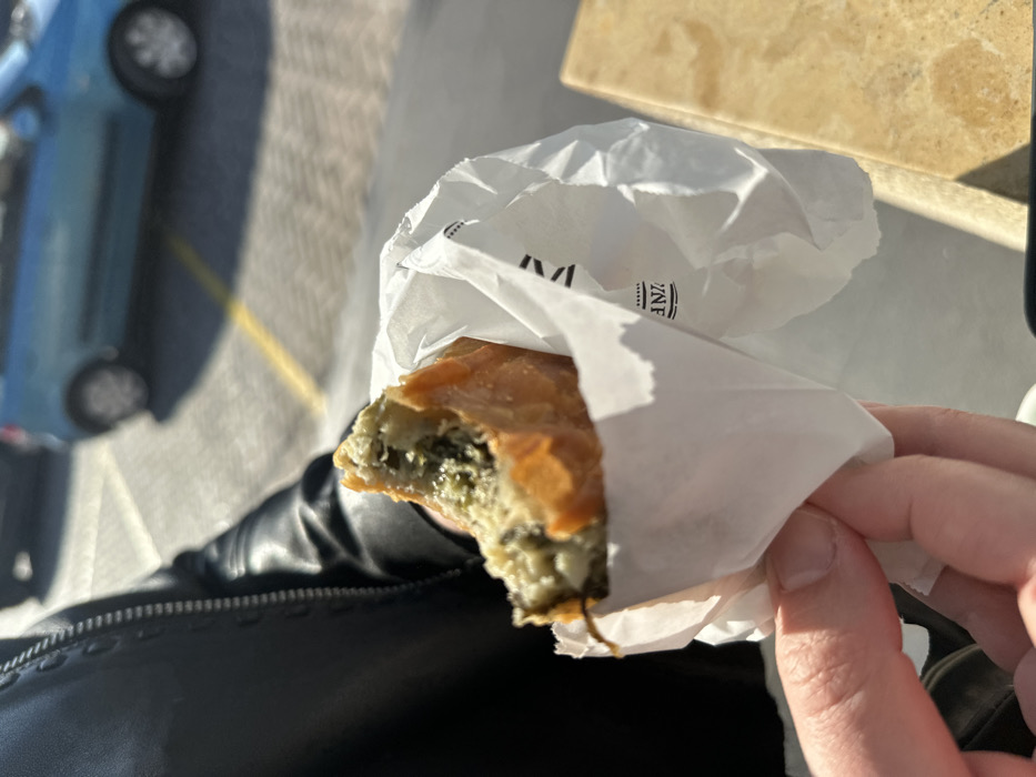 The image presents a first-person perspective, focusing on a hand holding a partially consumed savory pastry, likely a spanakopita, wrapped in white paper. The pastry features a golden, flaky crust, revealing a vibrant green filling, indicative of spinach and possibly cheese. The person's fingers are visible, along with a portion of a black sleeve or jacket.
The white paper wrapper displays a printed logo. The word "ZINE" is clearly visible, with less distinct text above it, possibly "VIN" or "VI," and smaller, blurred text beneath it, which could be a descriptor like "BAKERY" or "PATISSERIE." The logo is accented with small dots, suggesting a brand for a food establishment.
In the background, a light blue car is parked on a paved surface, out of focus. To the upper right, a blurred, light-colored surface, possibly marbled stone, suggests a table or counter. The scene is illuminated by strong, natural sunlight, indicating daytime. The overall setting appears to be outdoors or semi-outdoors, capturing a casual moment of eating. This activity is taking place in Metamorfosi, Greece.
