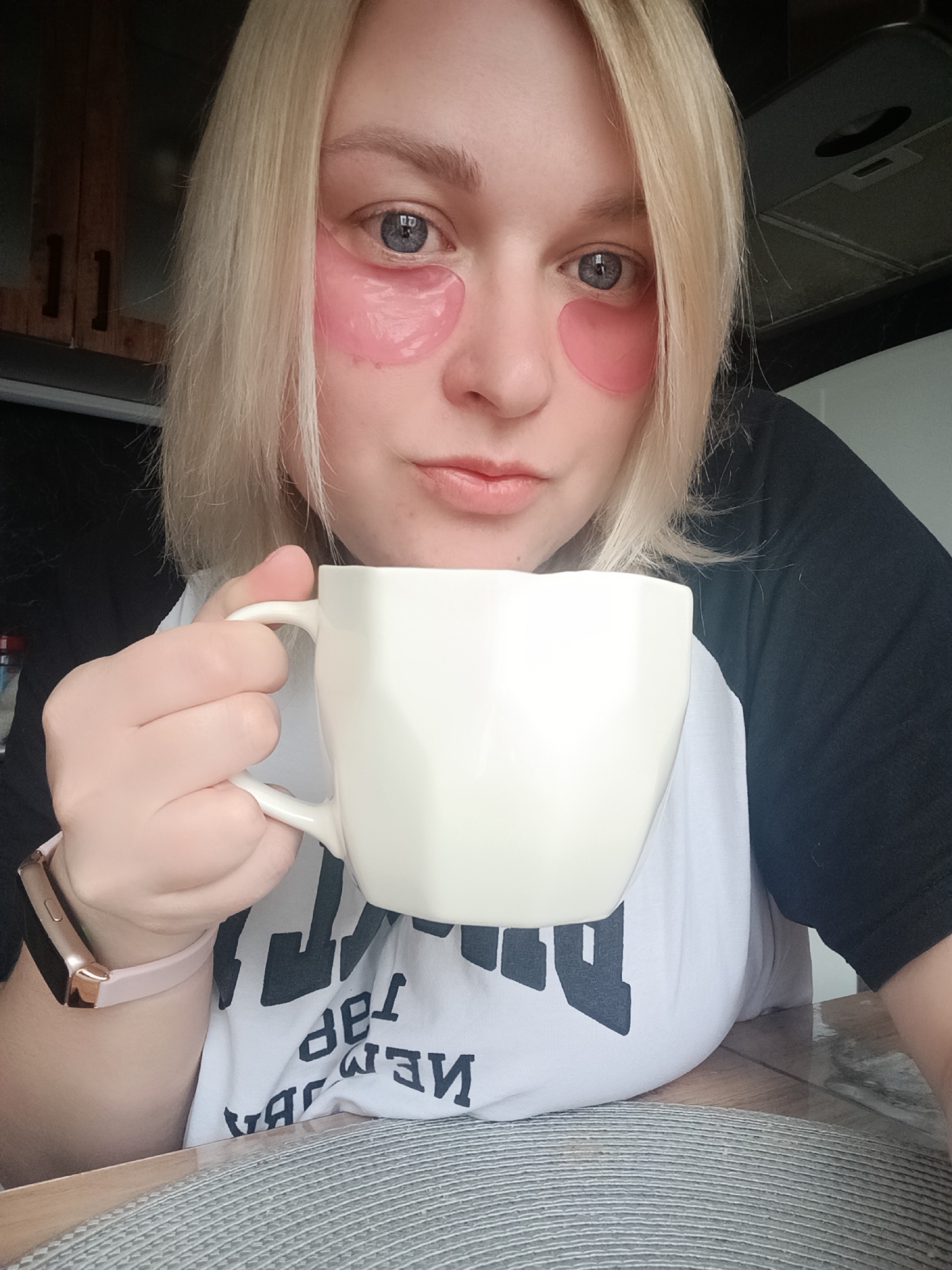 A medium close-up shot captures a blonde woman from the chest up, engaged in a relaxed indoor activity. She has shoulder-length, light blonde hair that frames her face, and her blue-grey eyes gaze directly at the camera. On her cheeks, beneath each eye, she wears pink, gel-like under-eye patches, suggesting a skincare routine. Her expression is neutral, bordering on calm.
She is wearing a casual black and white raglan-style t-shirt. The white central part of the shirt features inverted and partially obscured black text, which is difficult to decipher but appears to contain capital letters like "DeL" or "DeN". On her left wrist, a pink smartwatch with a rose gold-colored casing is visible.
The woman is holding a white, geometrically faceted mug with both hands, positioned in front of her. The setting appears to be a home kitchen or dining area. In the background to the left, wooden kitchen cabinets with metal handles are visible. To the right, a dark kitchen exhaust hood or ventilation system is mounted overhead. The woman is leaning on a shiny surface, possibly a table or countertop, which is covered by a gray, textured placemat beneath her hands. The lighting suggests it is daytime. The overall scene conveys a moment of a casual morning or daytime routine, possibly in Chaikovsky, Russia.