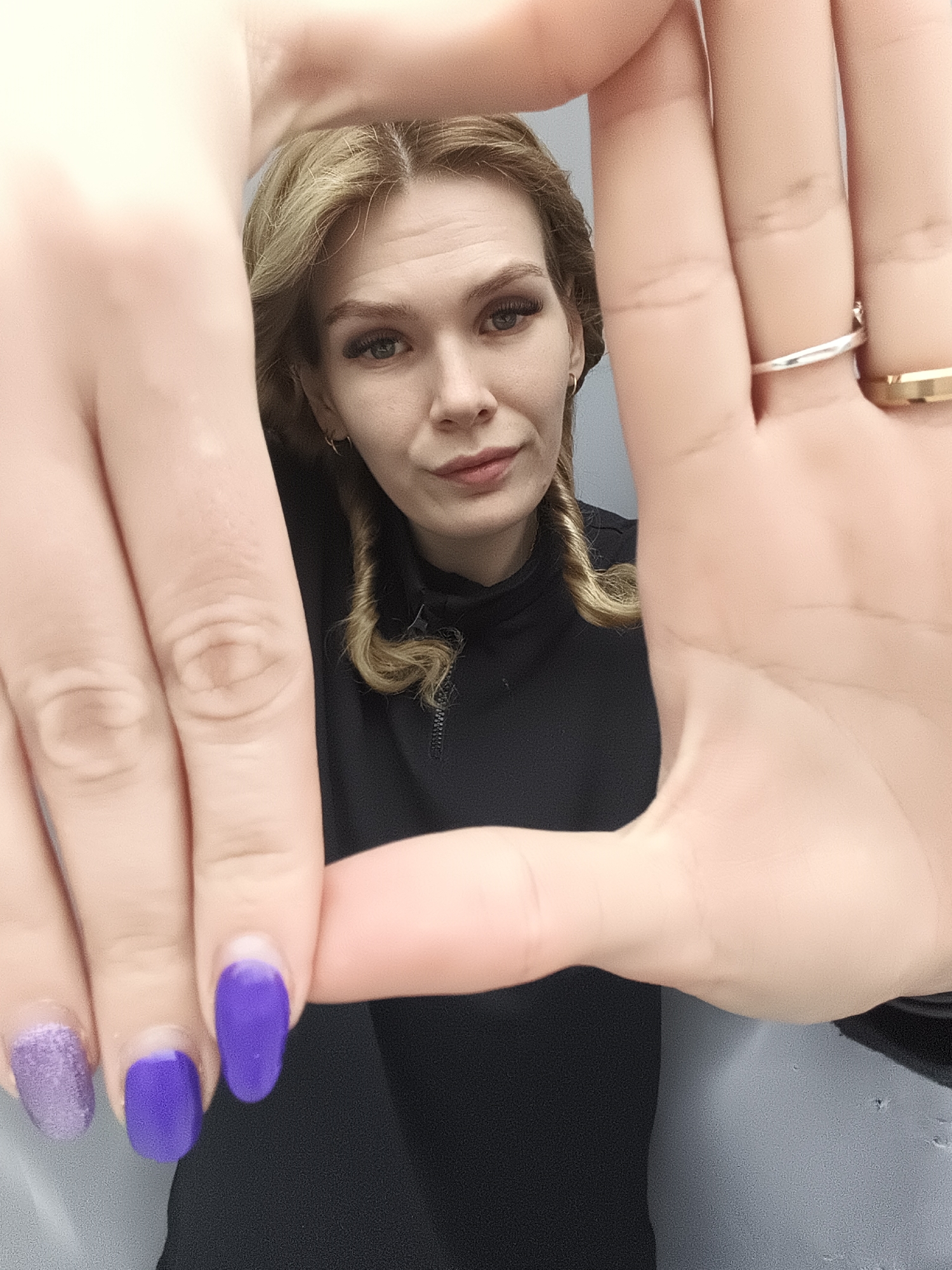 The central subject of the image is a woman with blonde or light brown, wavy hair, framed by her hands in the foreground. Her gaze is directed towards the viewer. She has blue eyes and a subtle, serious or contemplative expression with a slight hint of a smirk on her lips. Her hair features distinct curly tendrils on the sides.
Her hands are prominent, creating a "frame" around her face. Two rings are visible on her right hand: a thin silver band on her ring finger and a wider, gold-toned band on her pinky finger. Her fingernails are painted in shades of purple; some are a solid vibrant purple, and one has purple glitter. There are visible signs of nail regrowth at the base of the nails.
The woman is wearing a black, high-necked top with a partial zipper. The background appears to be an indoor setting, with a light-colored area behind her head transitioning to a darker, possibly grey, surface on her right.
The activity taking place is the woman posing for a photograph, intentionally using her hands to frame her face. No specific time of day or weather is discernible from the image. The location context provided is Русский Вожой, Russia. There is no visible text in the image.