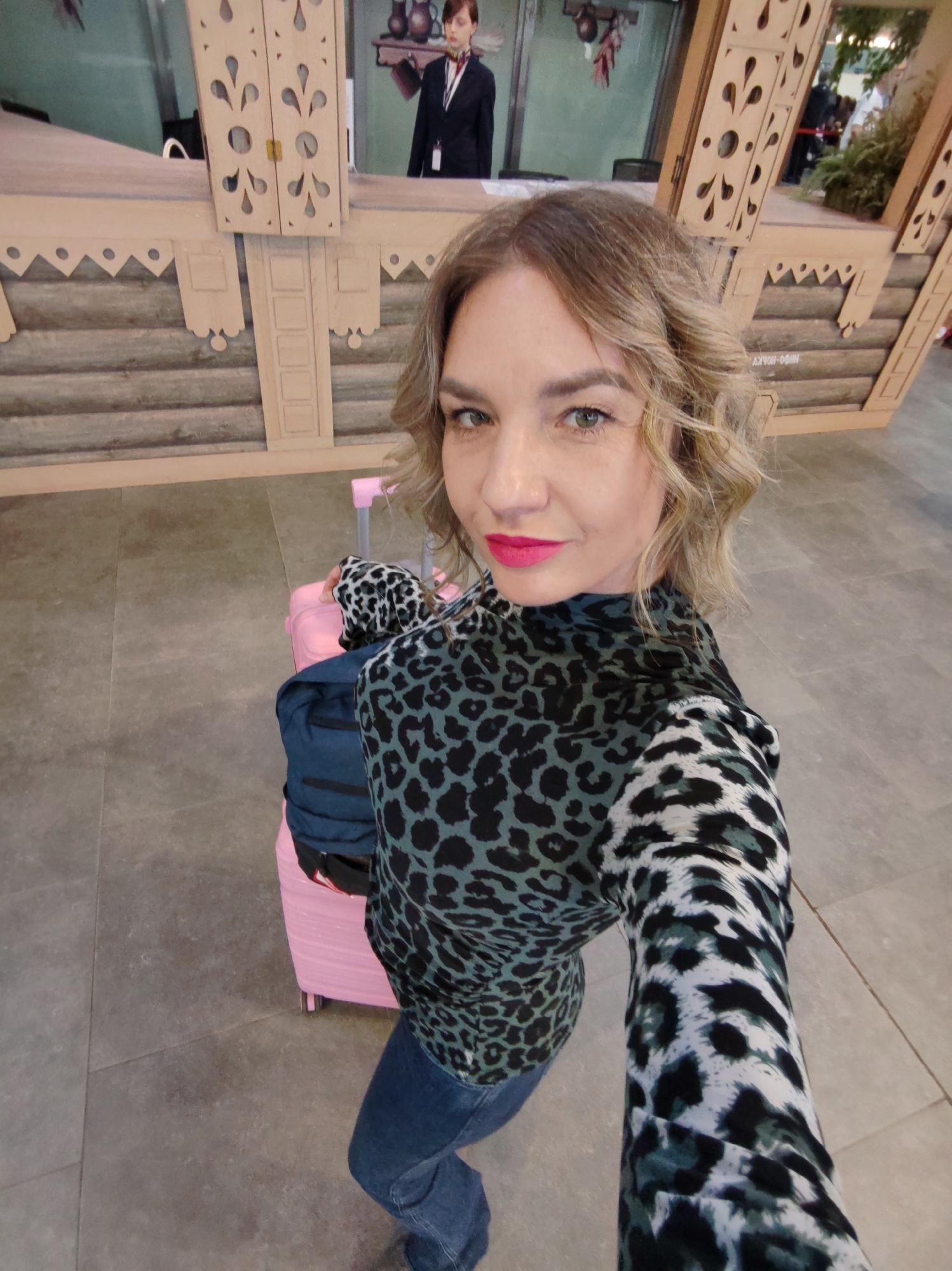 A woman takes a selfie indoors, looking directly at the camera with a slight smile and wearing bright pink lipstick. Her hair is wavy and light brown. She is dressed in a long-sleeved top with a distinctive leopard print pattern, featuring both green/black and white/black sections, paired with blue jeans. Beside her, a pink wheeled suitcase and a blue backpack are visible, suggesting travel.
The background features an elaborate wooden structure, designed in a folk or traditional architectural style, resembling a stylized miniature log cabin or decorative booth with carved details. Behind this structure, another woman in a dark uniform stands behind a counter, appearing to be a staff member. The wall behind the counter has a mural or display with pottery and some green plants.
On the side of the wooden structure, the Cyrillic text "INFO-БЮРО" (Info Bureau) is visible, indicating an information desk and strongly suggesting a Russian location, consistent with the provided context of Saint Petersburg. The floor is covered with light grey tiles. The overall scene suggests an airport terminal, an exhibition, or a public event setting. The time of day is indeterminate but appears to be well-lit daytime.