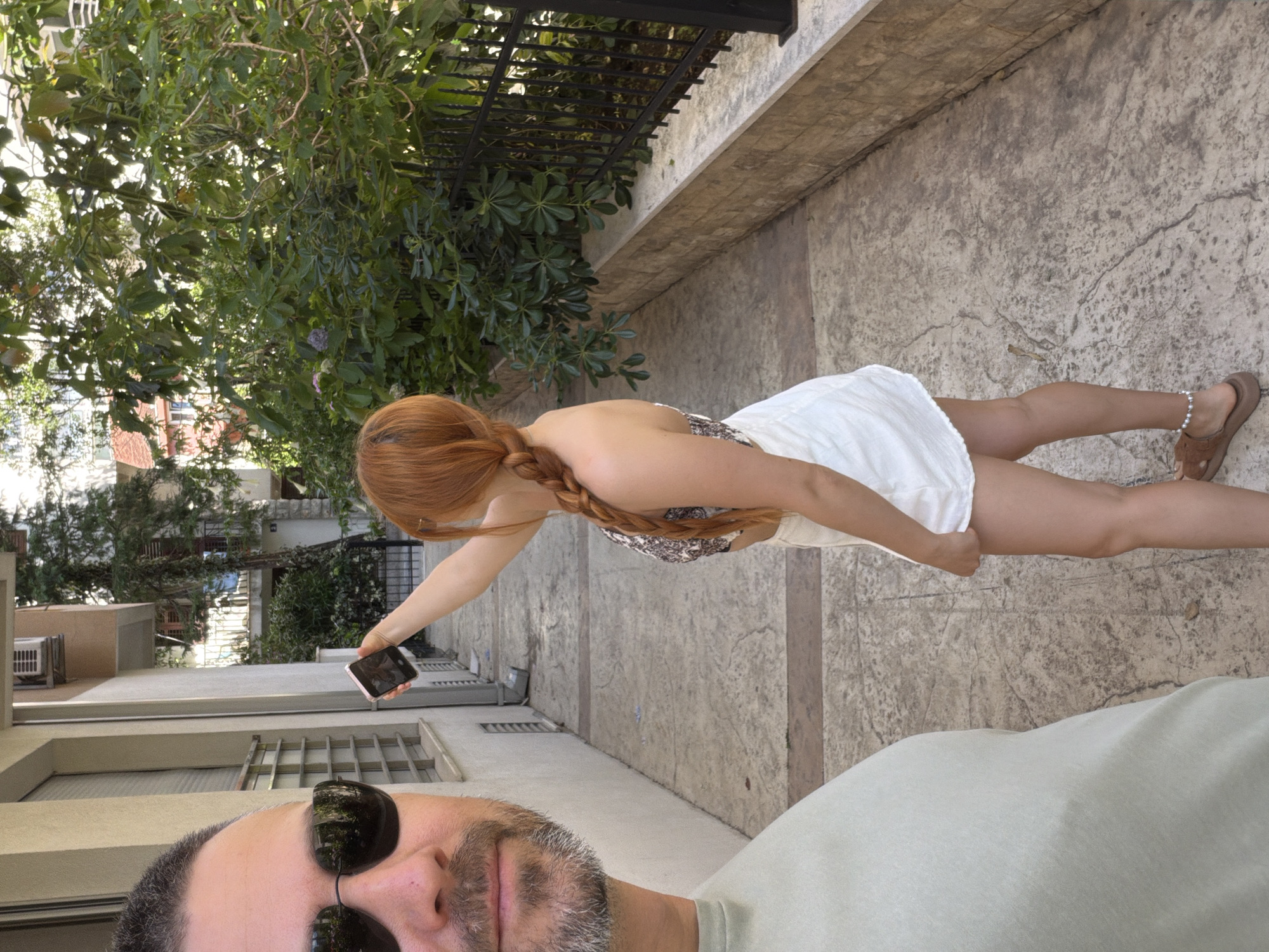 The image is a daytime, outdoor shot taken from a first-person perspective, likely a selfie. In the immediate foreground, the left side of a man's face and his upper torso are visible. He wears reflective dark sunglasses and has a neatly trimmed, graying beard. He is wearing a light green or khaki t-shirt.
Slightly further back and to the right, a woman with long, red hair styled in a thick braid stands facing away from the camera, slightly turned to her left. She wears a patterned, possibly animal print, halter-neck or backless top and white shorts. Her right arm is extended upwards, holding a light-colored smartphone vertically, with the screen facing away from the viewer, appearing to capture something above or to her side. On her right ankle, she wears a white pearl anklet, and brown open-toed sandals are visible on her feet.
The setting is a paved pathway with mottled grey and brown stamped concrete, featuring visible cracks. To the left of the path, a light-colored building with several windows, an external metal grate, and an air conditioning unit mounted high on the wall is present. A grey downspout runs vertically down the building. To the right, a low stone wall made of stacked light brown blocks supports a black metal fence with vertical bars. Behind the fence and wall, lush green bushes and trees, some with small purple flowers, are visible. Through the foliage, other buildings with balconies and one with a distinctive reddish-orange tiled roof can be seen in the background. The scene is brightly lit by natural sunlight, indicating daytime in the city of Budva, Montenegro.
