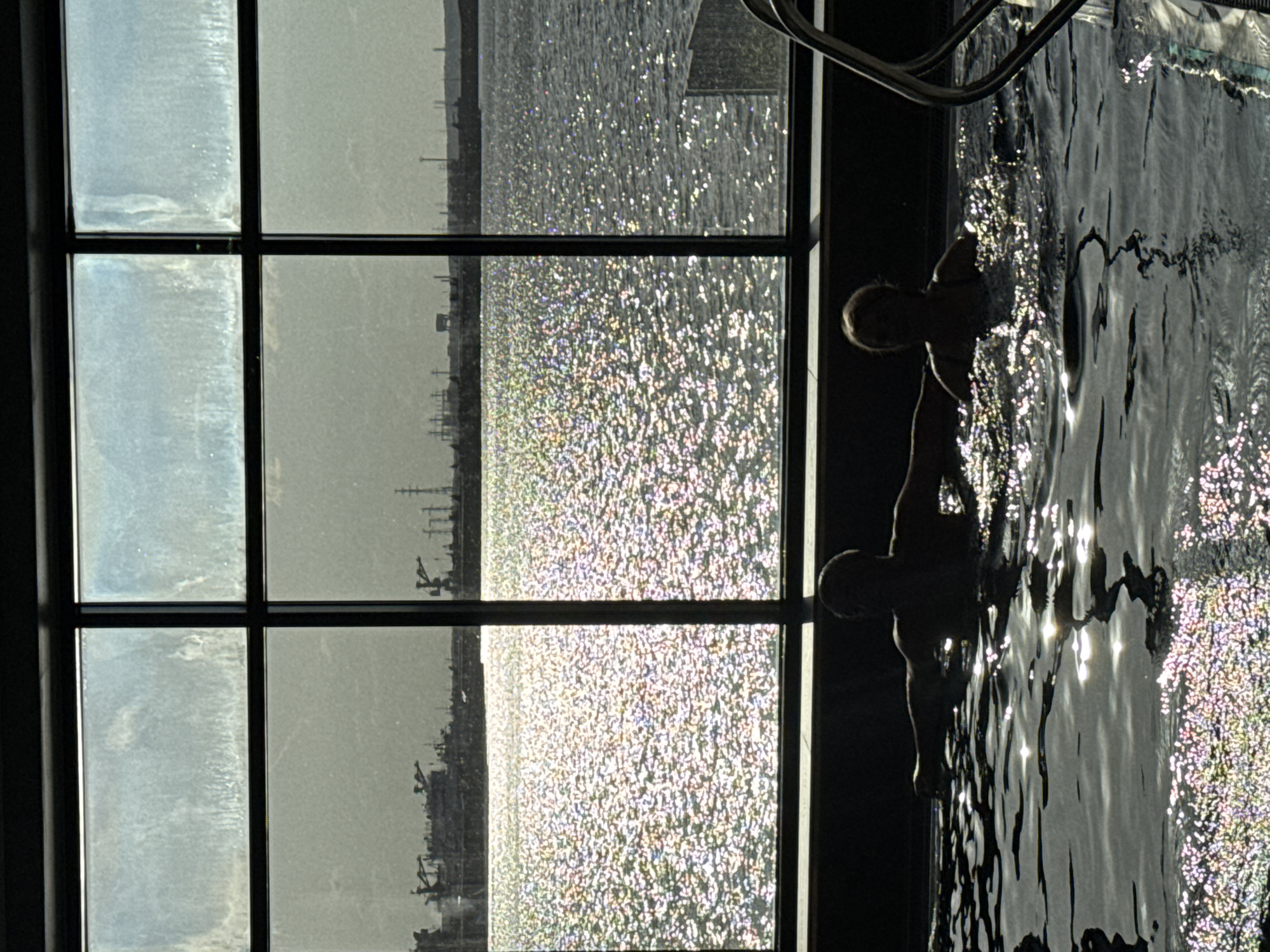 A view from an indoor swimming pool is captured through a gridded window. The window looks out onto a body of water, likely a river or lake, with land and structures visible in the distance. Sunlight reflects off the water's surface, creating a shimmering, speckled effect. Two people are visible in the water; their bodies are partially submerged. A metal ladder can be seen at the pool's edge. The reflection of the sunlight is visible on the surface of the water in the pool. The location is likely Chaikovsky, Russia, based on the image's origin.