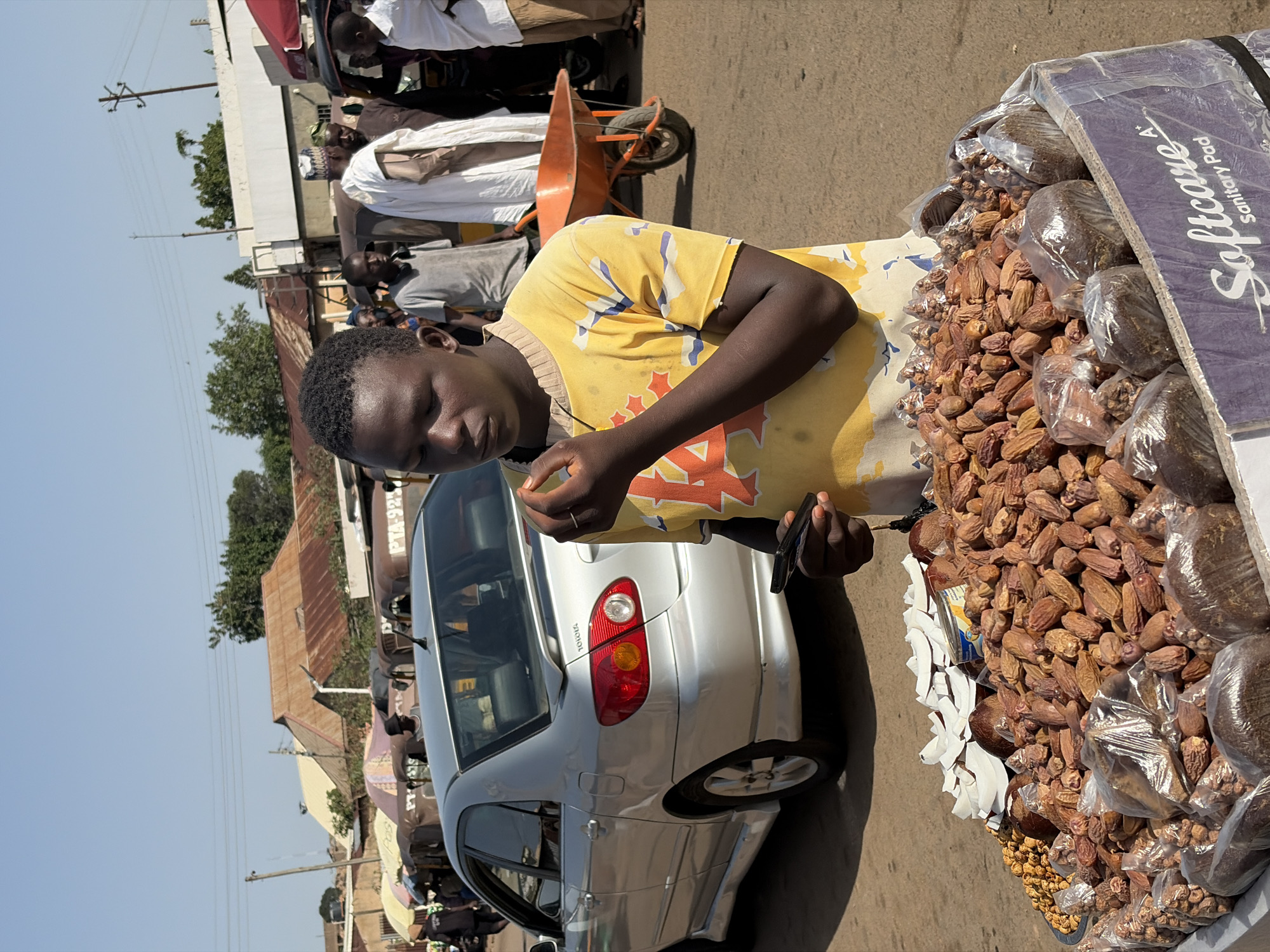 The image shows a street scene in Gura Top, Nigeria. A man in a yellow patterned shirt stands next to a silver Toyota Corolla. The car is parked close to a table laden with dates and packaged goods for sale. The man is holding an object, possibly food, in one hand and a dark-colored device, possibly a phone, in the other. In the background, other people are visible, and there's a wheelbarrow nearby. Buildings and utility poles are also visible in the background. The lighting suggests it is daytime.