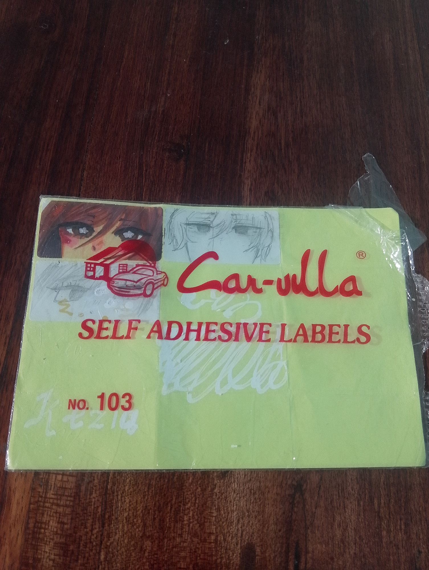 The image shows a light green package of "SELF ADHESIVE LABELS" by "Car-ulla" with the number "NO. 103" printed on it. The package is partially covered by a clear plastic wrap. On the top left of the package, there are drawings of anime-style eyes and a house with a car in front of it. On the right side of the package, there are some scribbled drawings in white. The package is placed on a dark brown wooden surface. The location in the city of Serbelawan, Indonesia, is not specified in the image.