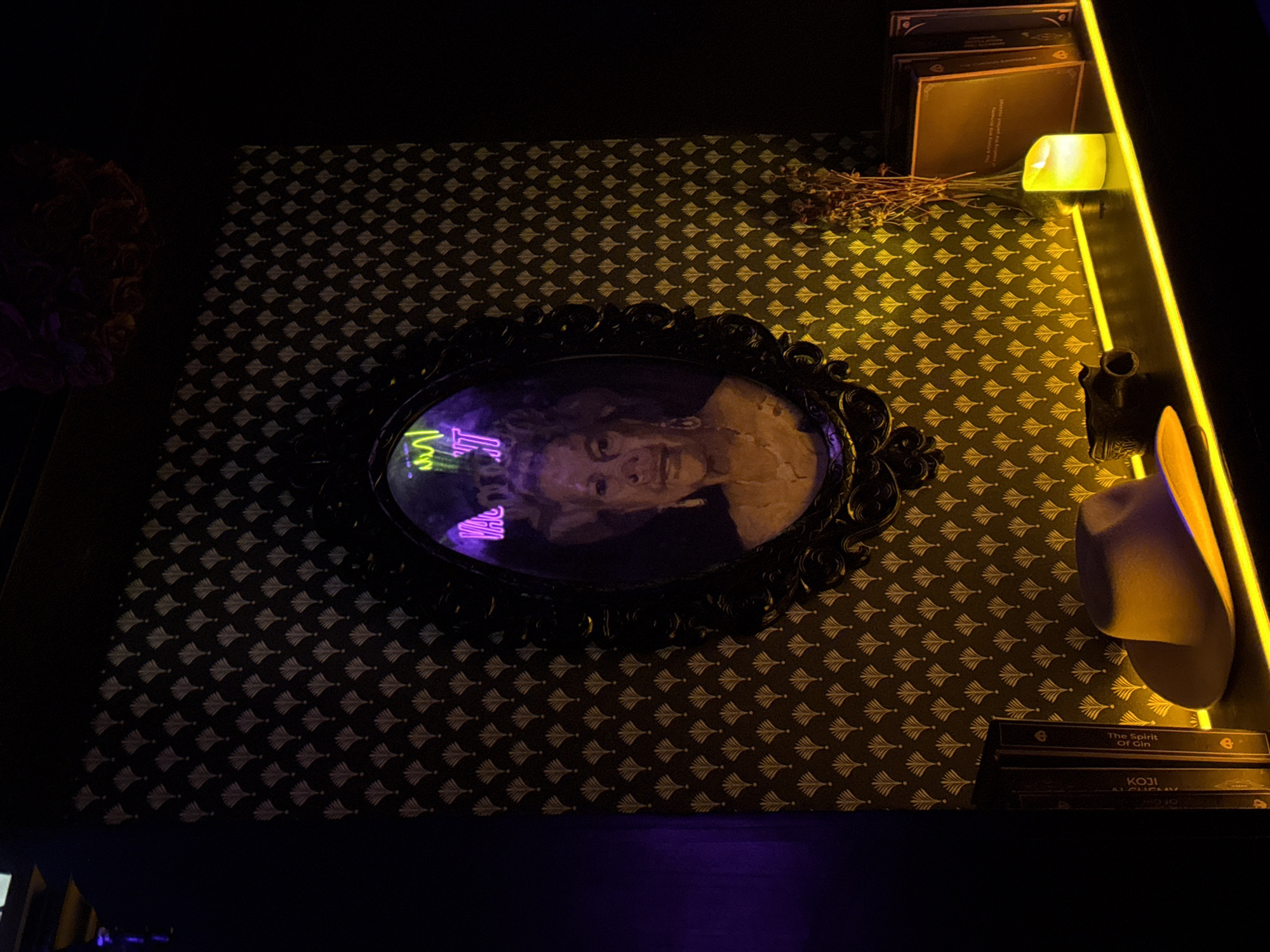 This is a photograph taken in North Kuta, Indonesia, depicting a dimly lit interior scene. A portrait of an older woman hangs in an ornate, dark frame against a dark wallpaper with a repeating fan-like pattern in a lighter shade. A neon sign, partially visible and in shades of green and pink, is superimposed over the portrait.
In the foreground, a shelf is illuminated by a yellow neon strip. On the shelf are several objects: a light-colored fedora hat is placed on its side, and a pair of binoculars rests nearby. To the right, a vase with dried flowers stands next to a lit candle in a clear glass holder. Behind the candle, a framed object, possibly a book or artwork, is visible. Two books stand upright to the left of the hat, with some text visible on their spines. The overall atmosphere is moody and atmospheric, with the lighting creating strong contrasts.