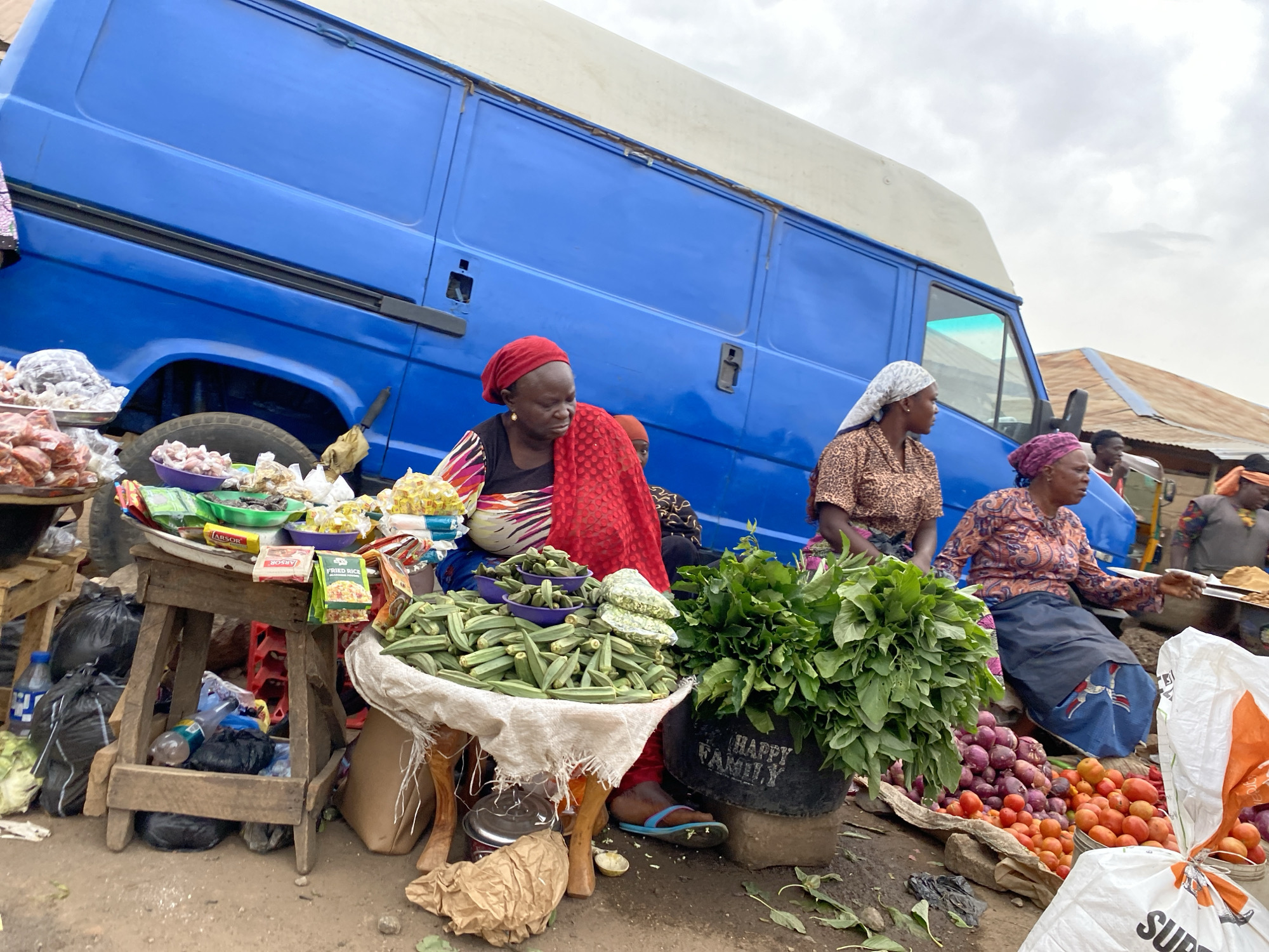 The image depicts a bustling roadside market scene in Bukuru, Nigeria. Several women are selling produce alongside a large blue van. One woman, wearing a red headscarf and patterned top, is seated behind a display of okra. Nearby, another woman in a patterned shirt and white headscarf attends to a pile of red onions and tomatoes. A third woman wearing a purple headscarf sits behind the produce, possibly assisting. Green leafy vegetables are displayed in a black bucket. Various goods are also on display on a makeshift wooden stand. Plastic bags, bottles, and other objects litter the ground. Other people are seen in the background, partially obscured. The sky is overcast.