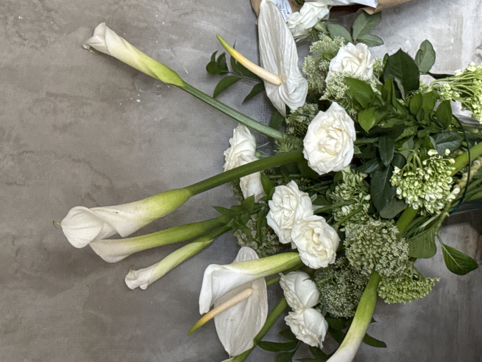 A bouquet of white flowers, including calla lilies and roses, is arranged on a gray concrete surface. The bouquet is positioned to the right of the frame, with several calla lilies extending towards the left. The roses and other green foliage form a denser cluster to the right. No people are visible. The scene appears to be indoors. North Kuta, Indonesia cannot be determined.