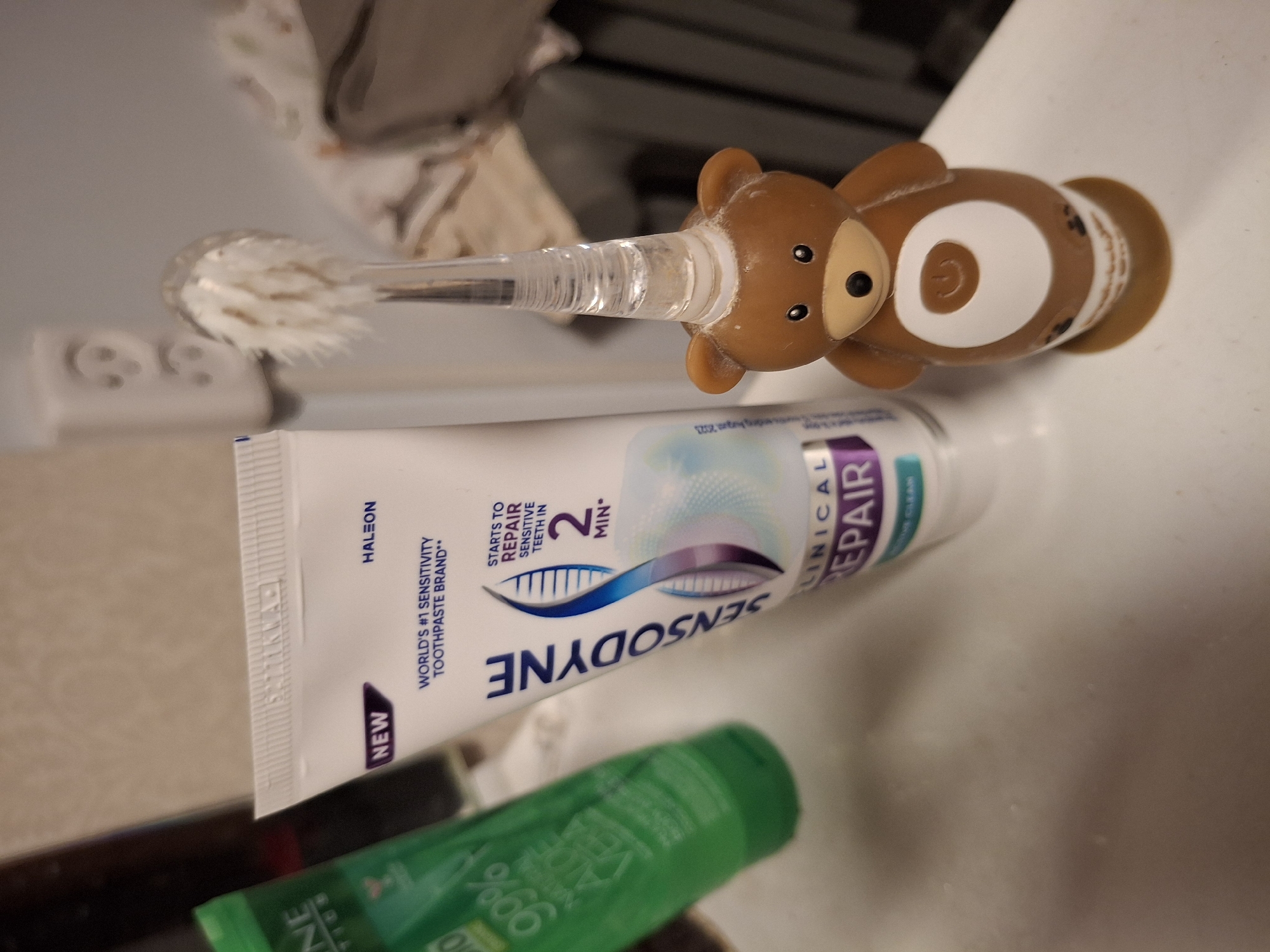 This is a close-up, horizontal shot of a bathroom sink area, with a focus on dental hygiene products. The primary subjects are a children's toothbrush with a teddy bear-shaped handle and a tube of Sensodyne toothpaste. The toothbrush has a clear plastic handle with a brown teddy bear head at the end, featuring small black eyes and a nose. The bristles of the toothbrush are visible and appear slightly used. The tube of toothpaste is white with blue and purple accents and text. Visible text on the toothpaste tube includes "HALEON," "NEW," "WORLD'S #1 SENSITIVITY TOOTHPASTE BRAND," "SENSODYNE," and "CLINICAL REPAIR." There are also mentions of "STARTS TO REPAIR SENSITIVE TEETH IN 2 MIN." A partially visible green bottle, likely another personal care product, is in the foreground. The background is slightly blurred, showing what appears to be a tiled surface or countertop and possibly fabric. The lighting suggests an indoor setting, likely during the day given the overall brightness. There is no indication of people or a specific event, but the items suggest routine personal hygiene. The location context provided is "Ogre, Latvia," but this information is not visually evident from the image itself.