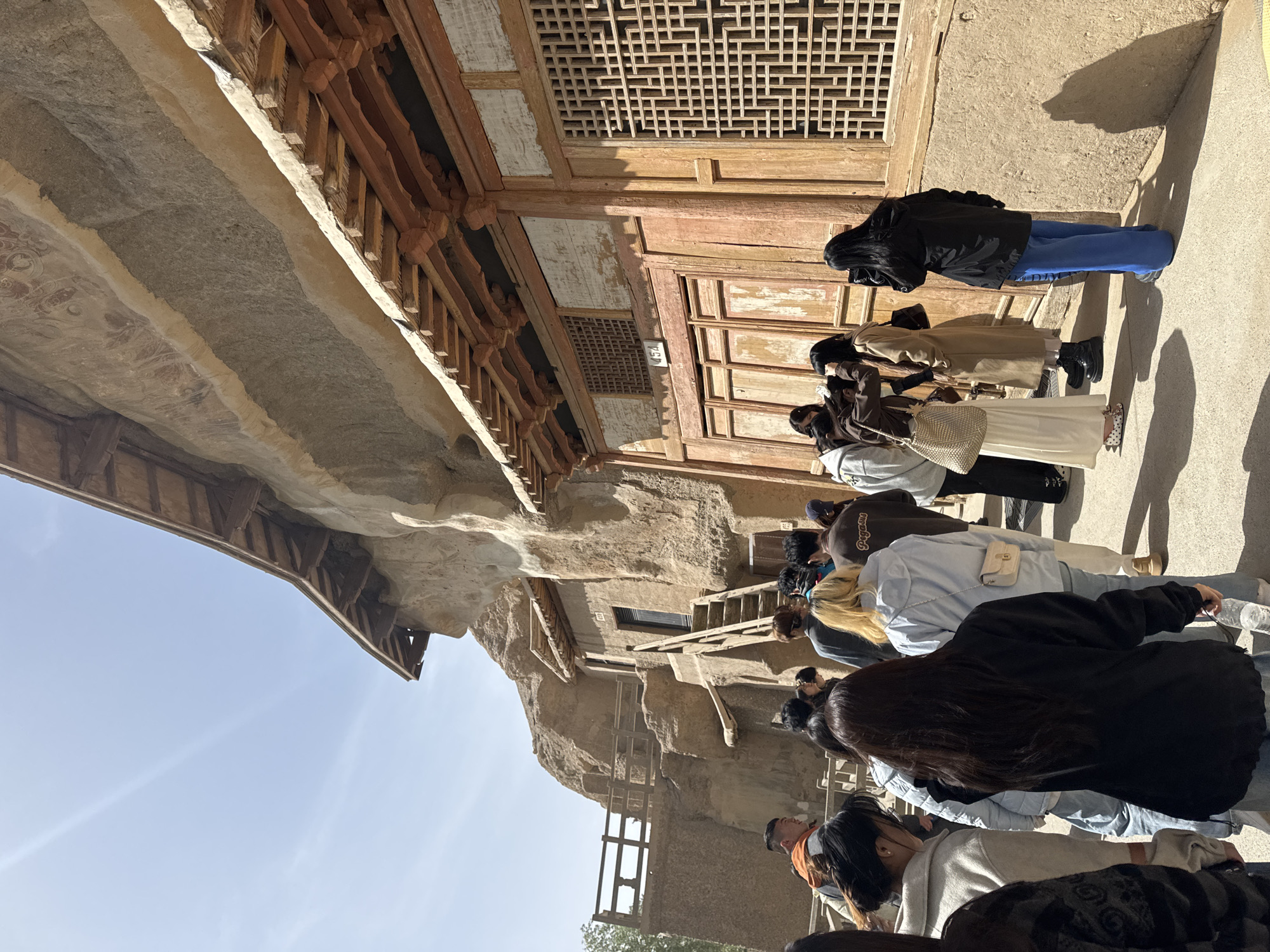 The image shows a group of people, presumably tourists, standing in a line in front of a historic building in Dunhuang City, China. The building appears to be a part of the Mogao Caves complex, judging from its architectural style featuring wooden lattice windows, exposed rock formations, and painted ceilings. The architecture is a blend of wooden structure and cave-like construction. The tourists are facing the building, suggesting they are looking at and learning about the structure. They are wearing casual clothing. The sky is visible and indicates a clear day.