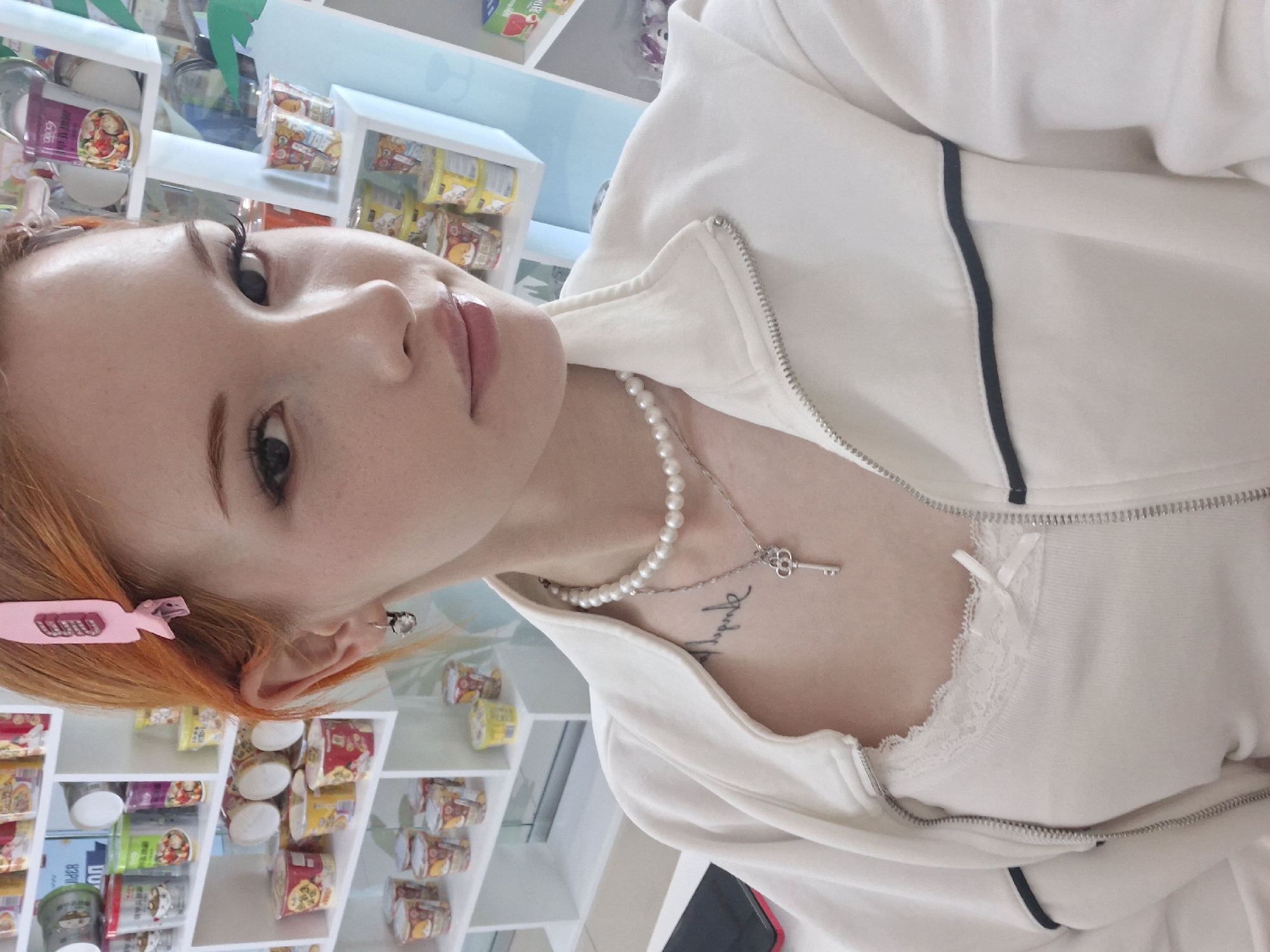 A young fair-skinned woman with strawberry-blonde hair poses for a close-up photo. A pink hair clip is in her hair. She wears a pearl necklace with a silver key pendant and a white zip-up jacket with black trim. Part of a white lace garment is visible under the jacket. The background includes white shelves stocked with food containers. This photograph appears to be taken indoors, possibly in a store or residence in Chaikovsky, Russia, judging by the request context.