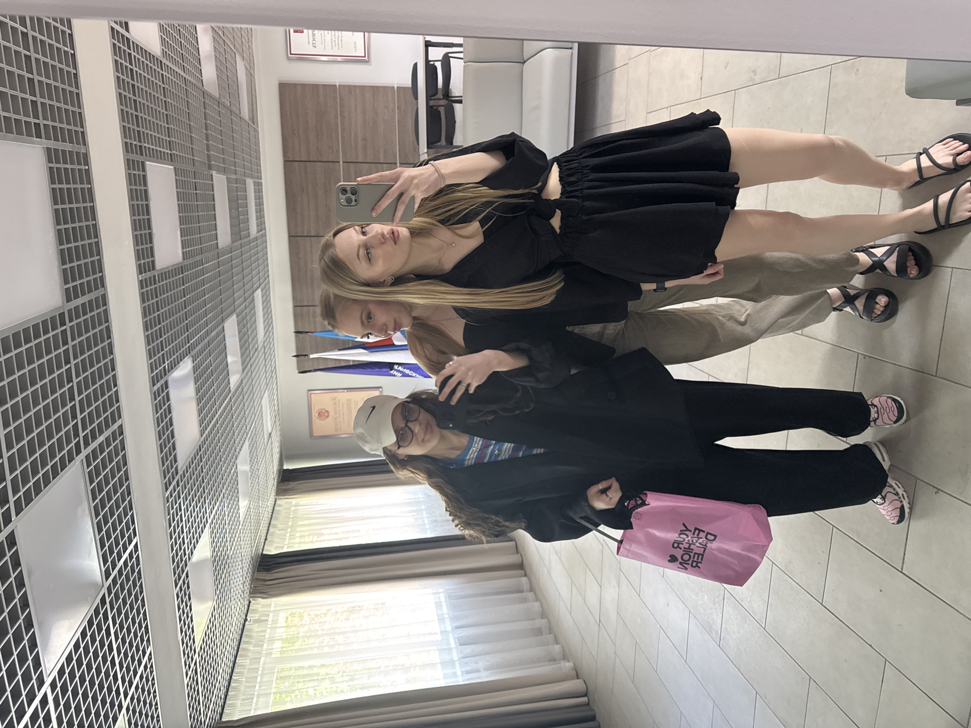 Three women are posing for a photo in what appears to be a reflection in Perm, Russia. The woman in the foreground wears a black blazer, black pants, and pink sneakers with a pink shopping bag. The woman in the middle is wearing tan pants. The woman in the background is wearing a black dress. They are in a room with a gray and white patterned wall, white curtains, and a white sofa in the background.