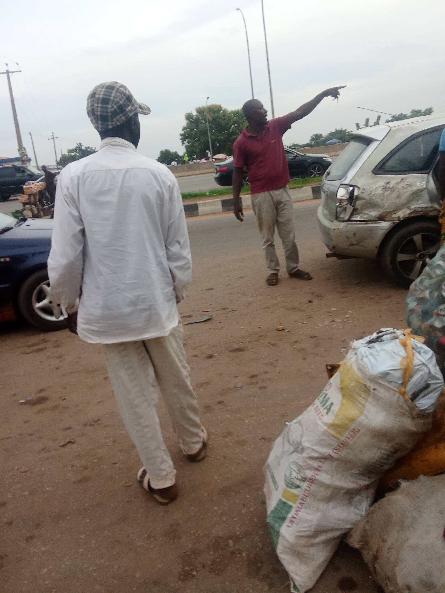 The image depicts a street scene in Kagini, Nigeria, with an apparent motor vehicle accident. In the foreground, a person wearing a white long-sleeved shirt and light-colored pants, along with a checkered cap, is observed from the back, facing away from the camera. To their right, another person in a maroon polo shirt and light-colored pants points towards the right side of the frame. Between these two individuals is a damaged silver car, showing significant impact to its rear section, including a caved-in bumper and crushed bodywork. The ground is unpaved, appearing to be a dirt or gravel road. Bags of what appear to be agricultural produce or supplies are visible near the damaged vehicle. In the background, other vehicles, trees, and utility poles are present, suggesting an urban or semi-urban environment. The sky is overcast.