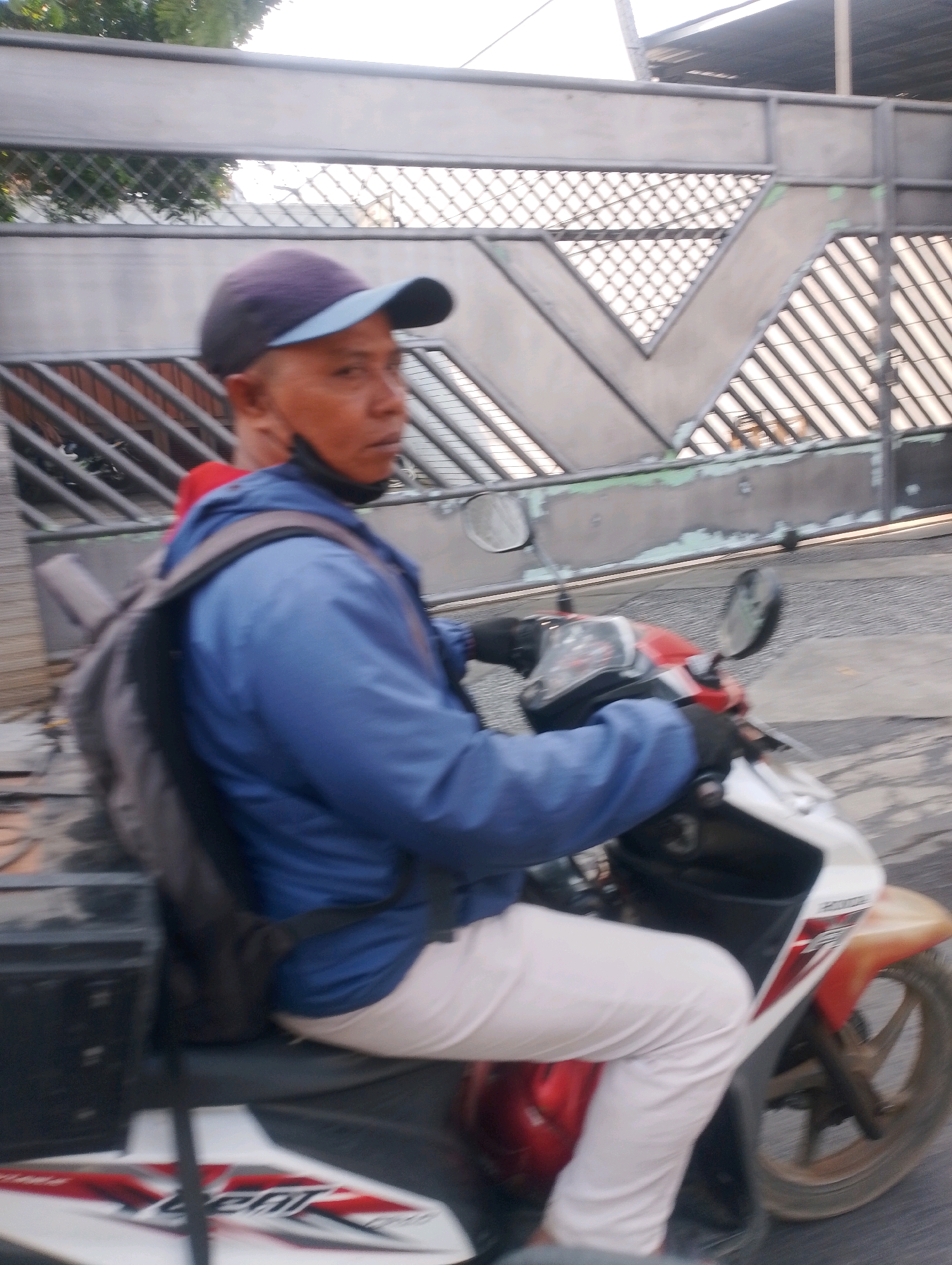 The image captures a side view of a person riding a motorcycle in what appears to be an urban environment in Probolinggo, Indonesia. The rider, wearing a blue jacket, light-colored pants, and a dark cap with a light blue brim, is seen from the chest down. A backpack is slung over their shoulders. The motorcycle is predominantly white with red and orange accents. The background features a metal gate with a diamond-shaped pattern and a diagonal bar design, suggesting it might be an entrance to a property or facility. The ground beneath the motorcycle appears to be asphalt or a similar paved surface. The lighting indicates it is daytime, with the sun possibly behind the viewer or casting shadows. The overall impression is that of a typical street scene in a residential or commercial area.